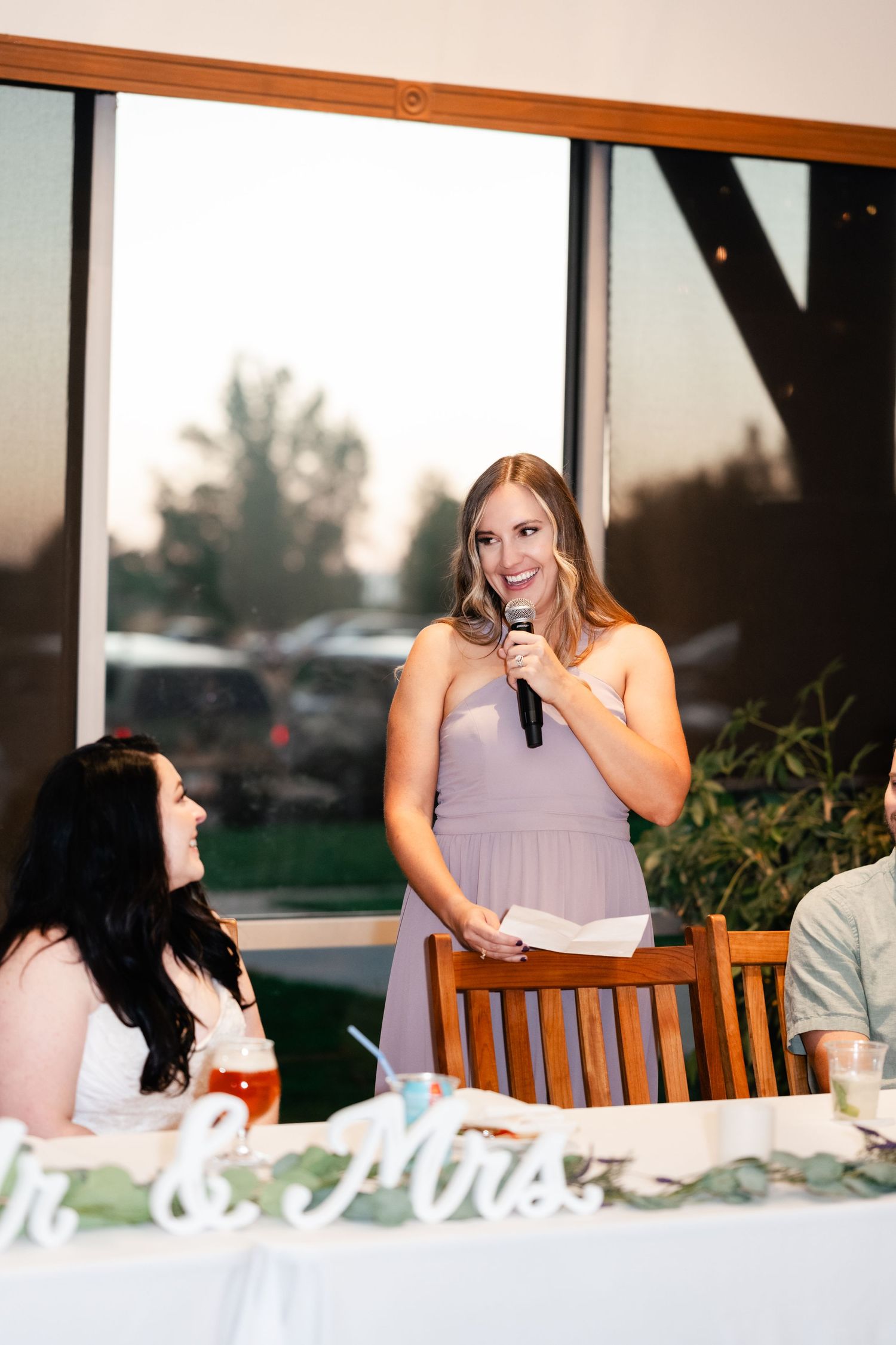 A wedding reception scene shows someone giving a speech at a table while holding a microphone during sunset.