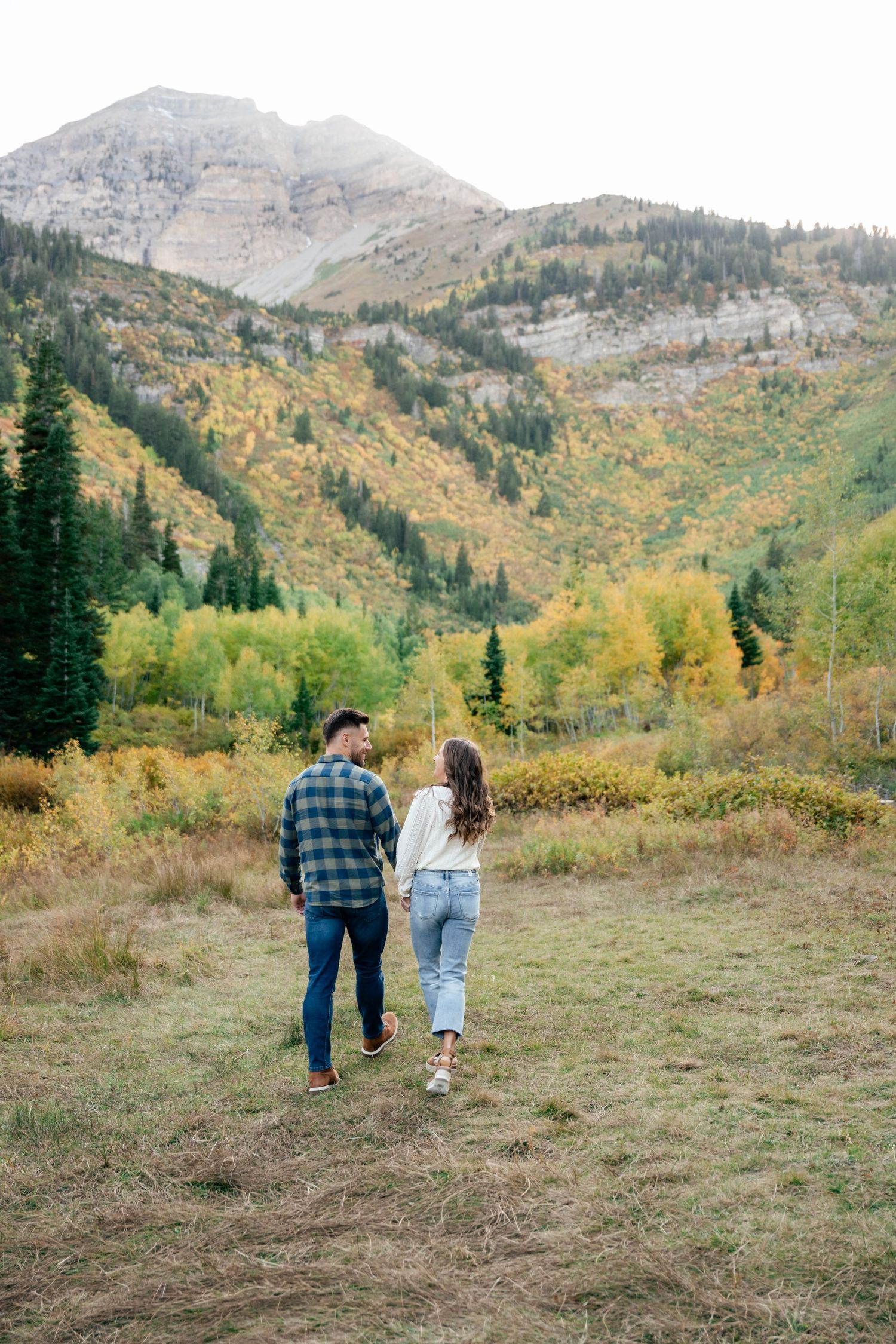 Two people walk hand in hand through a mountain meadow surrounded by autumn foliage and evergreen trees.