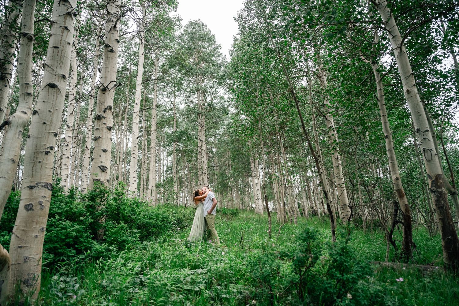 A couple stands together in a magical aspen forest with sunlight filtering through the white tree trunks. Park City engagement photographer