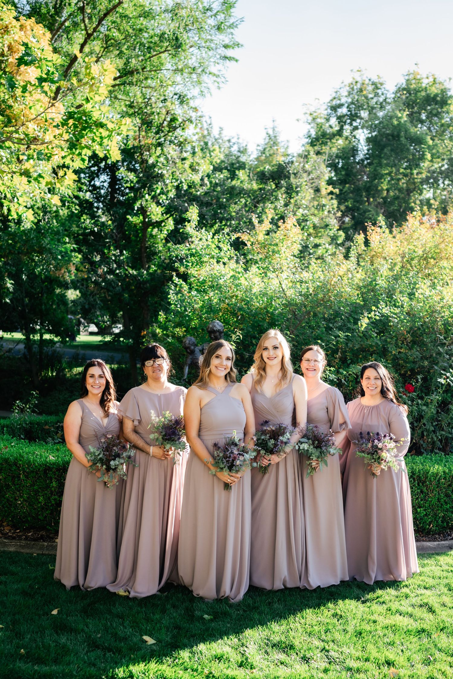 A group wearing matching blush-colored long dresses stands together on a lawn holding floral bouquets in a garden setting.