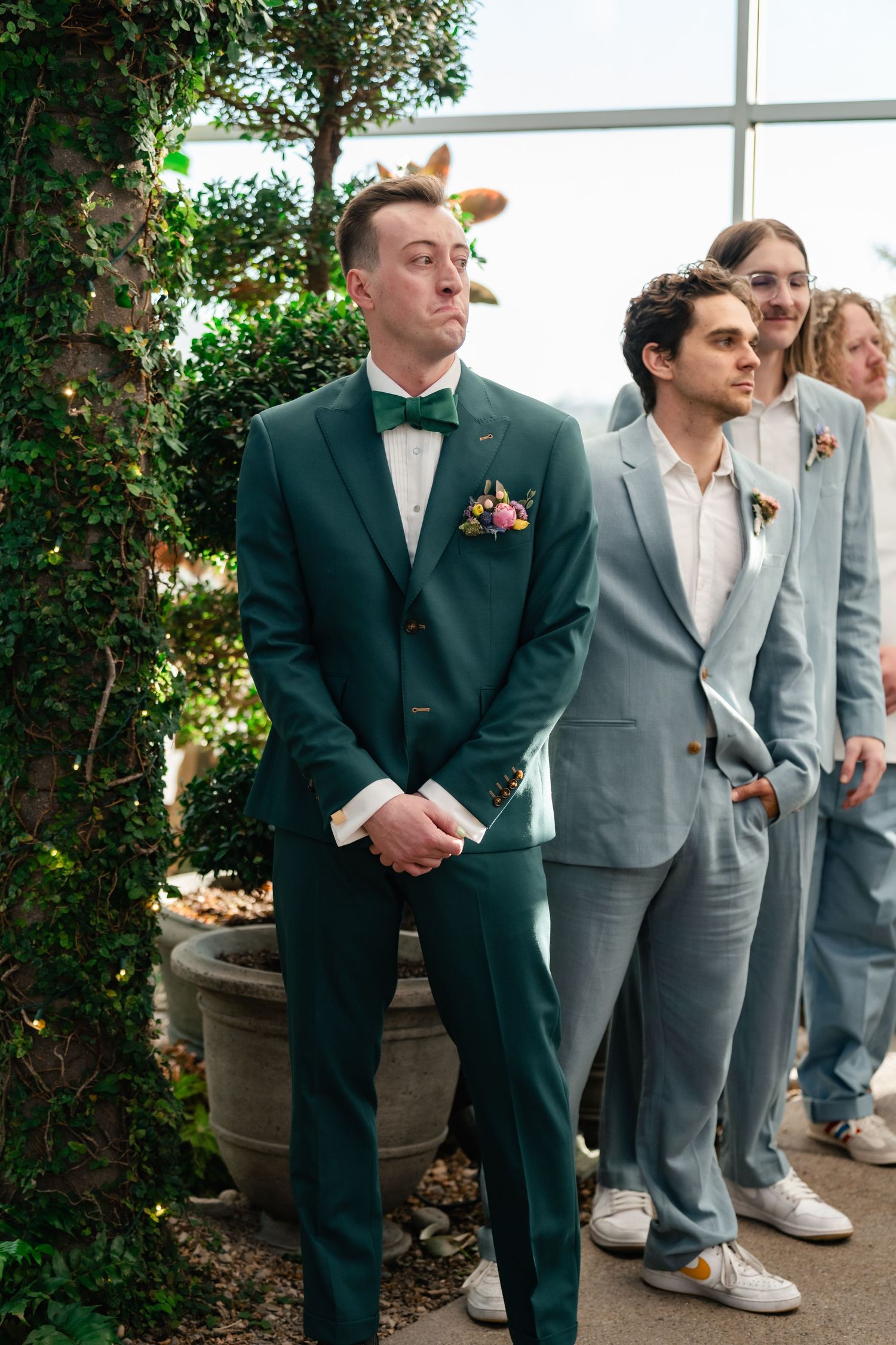 Wedding party groomsmen in dark and light suits stand together in a greenhouse setting.