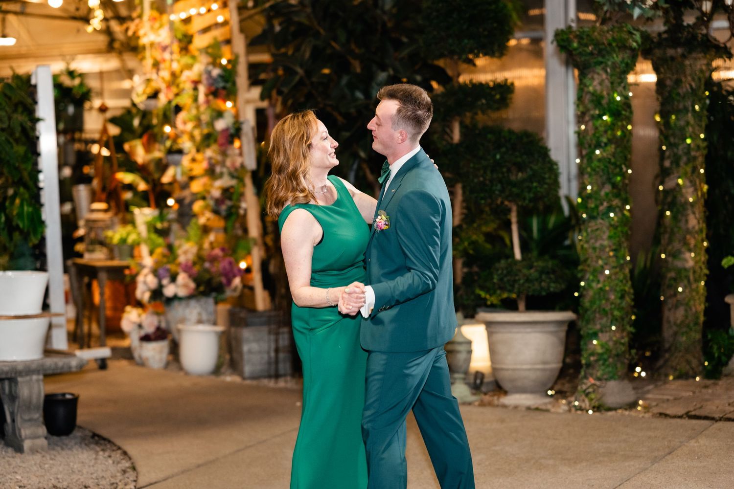 Couple dressed in matching emerald green formal attire dance together under twinkling lights in an outdoor setting.
