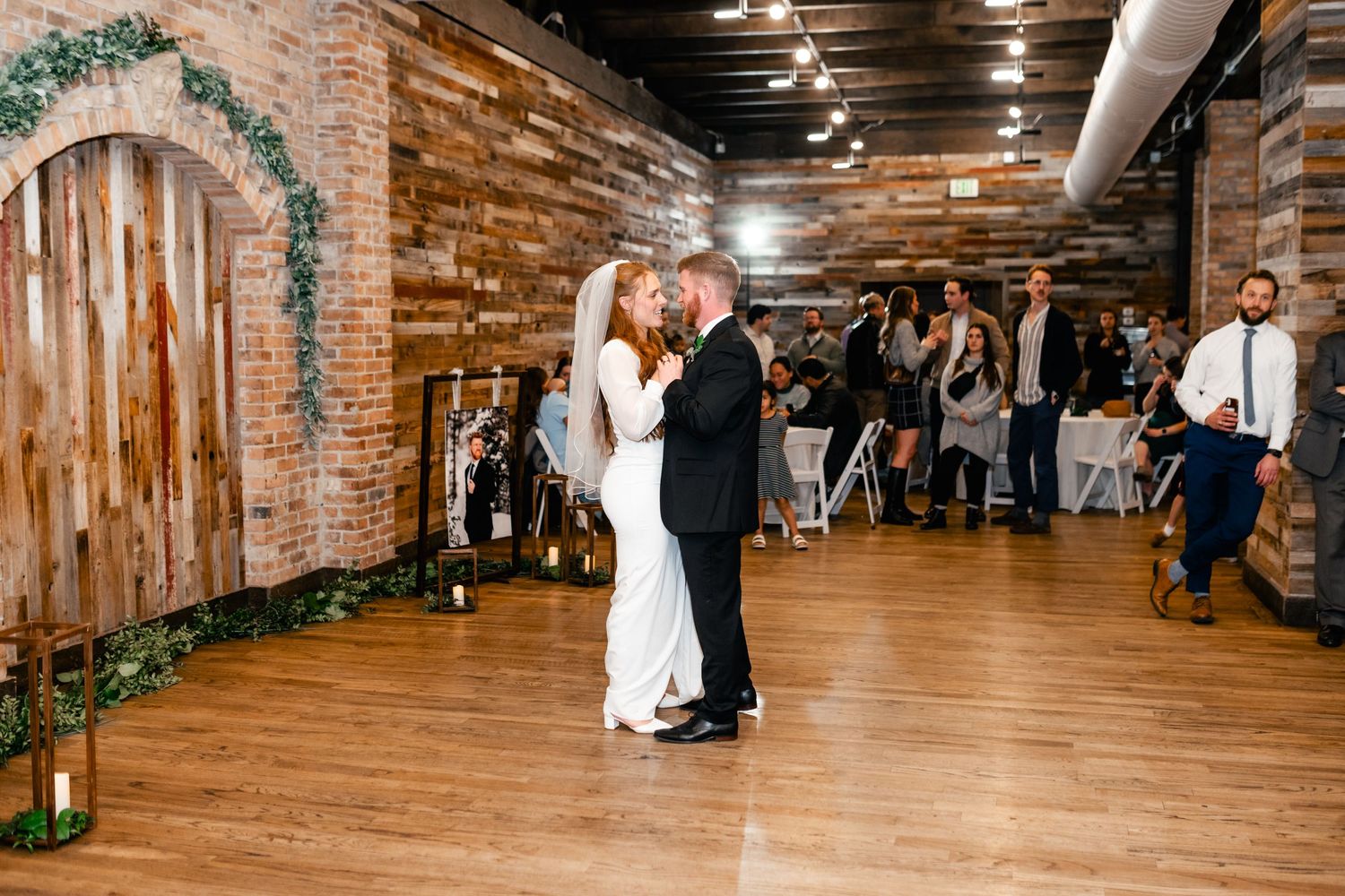 A couple shares their first dance in a rustic venue with exposed brick walls and hardwood floors while guests look on.