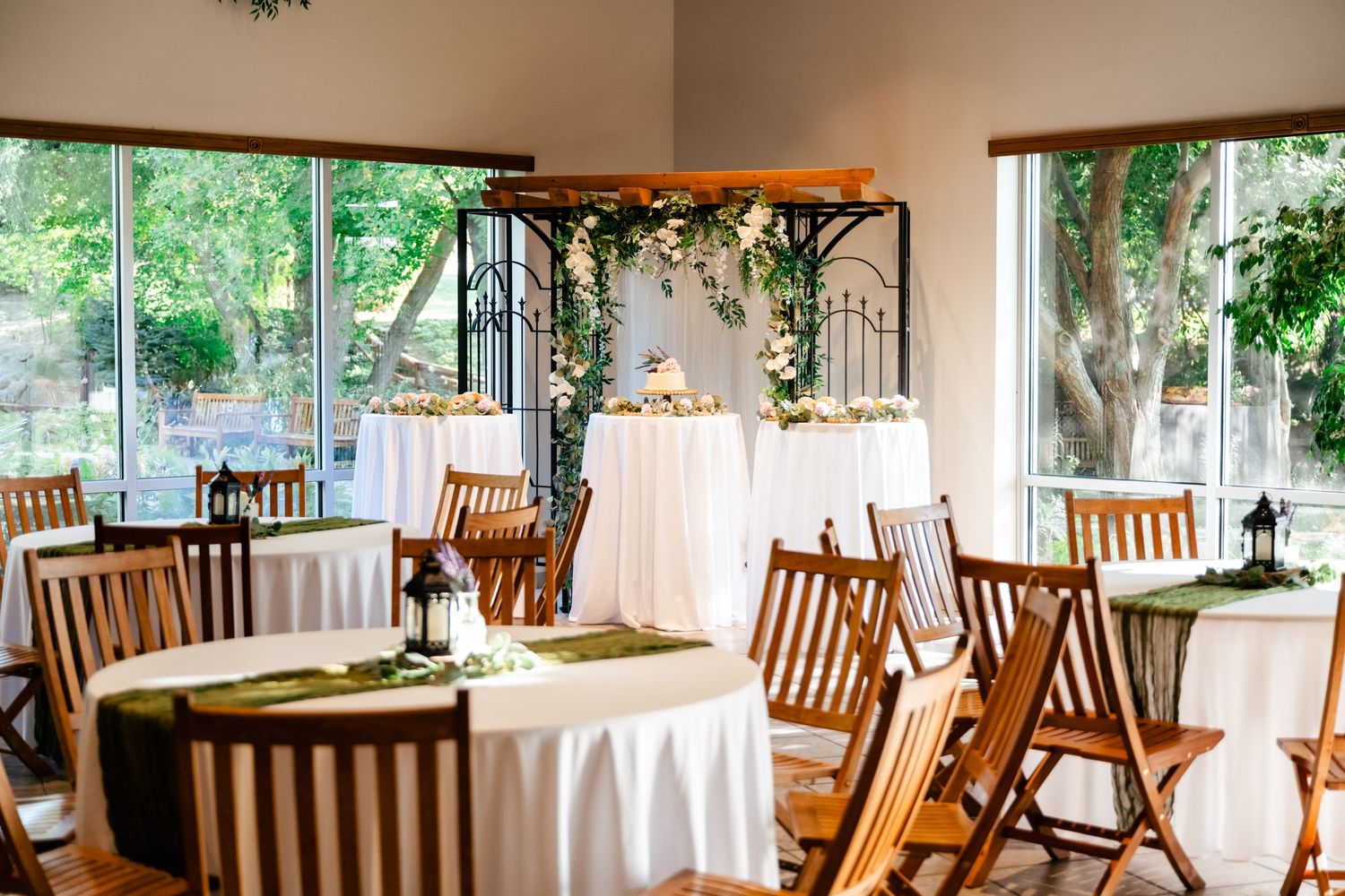A wedding reception venue with wooden chairs and round tables draped in white linens next to large windows with garden views.
