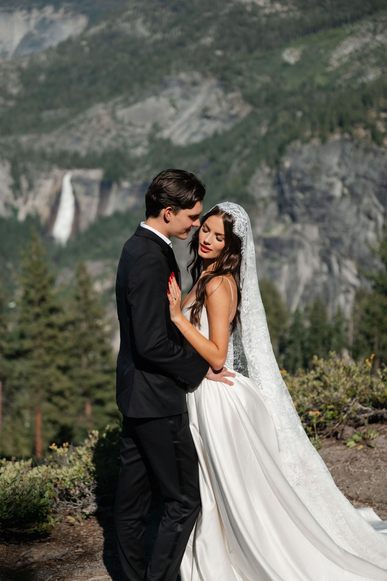 Bride and groom embrace at Yosemite with waterfall and mountain backdrop, bride in white gown with lace veil.