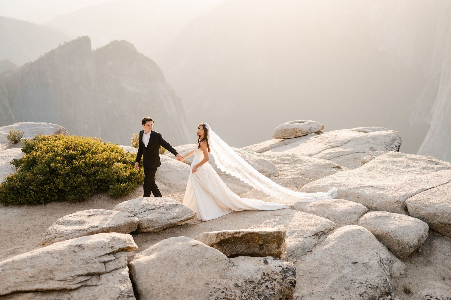 Bride and groom hold hands on rocky mountain summit with dramatic misty peaks, bride's veil flowing behind her.