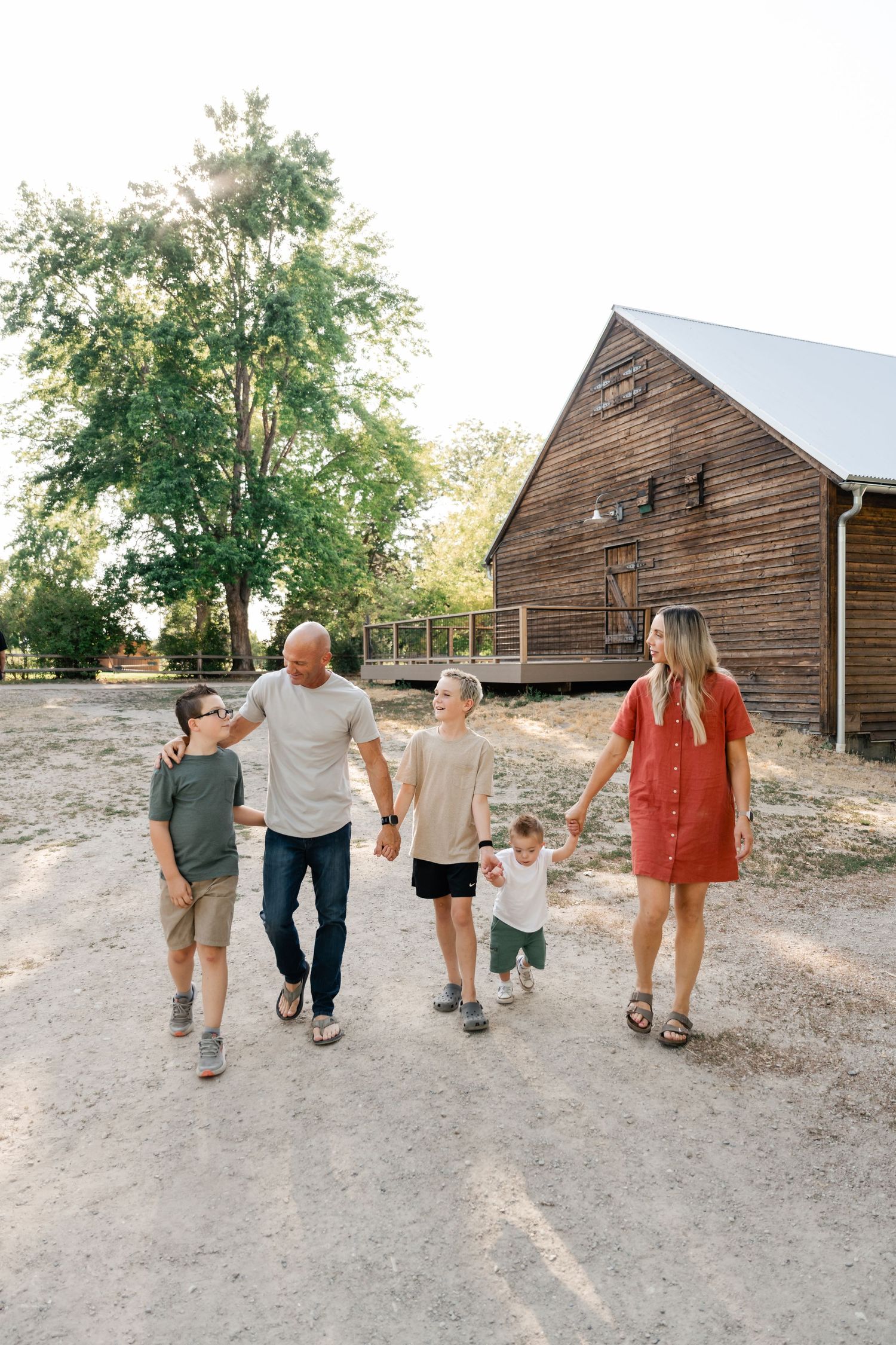 A family walks together on a dirt path with rustic wooden barns in the background during golden hour.