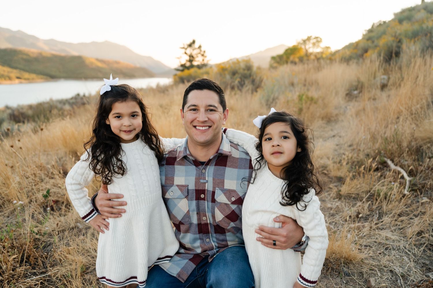 A family in casual attire poses together on a golden hillside during sunset with mountains in the background.
