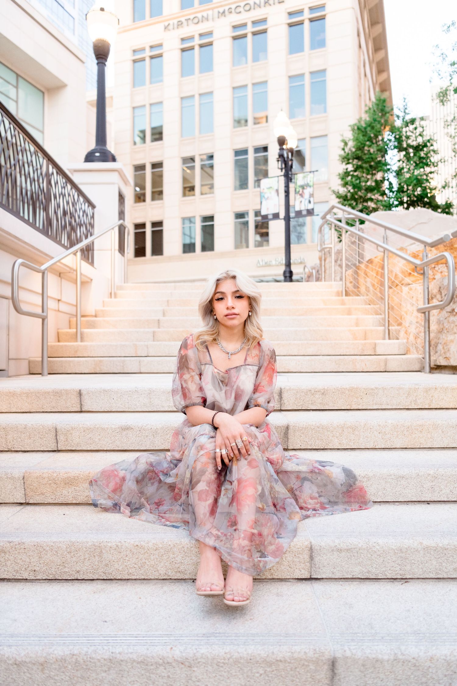 A person in a floral dress sits gracefully on white stone steps outside a building with elegant architecture.