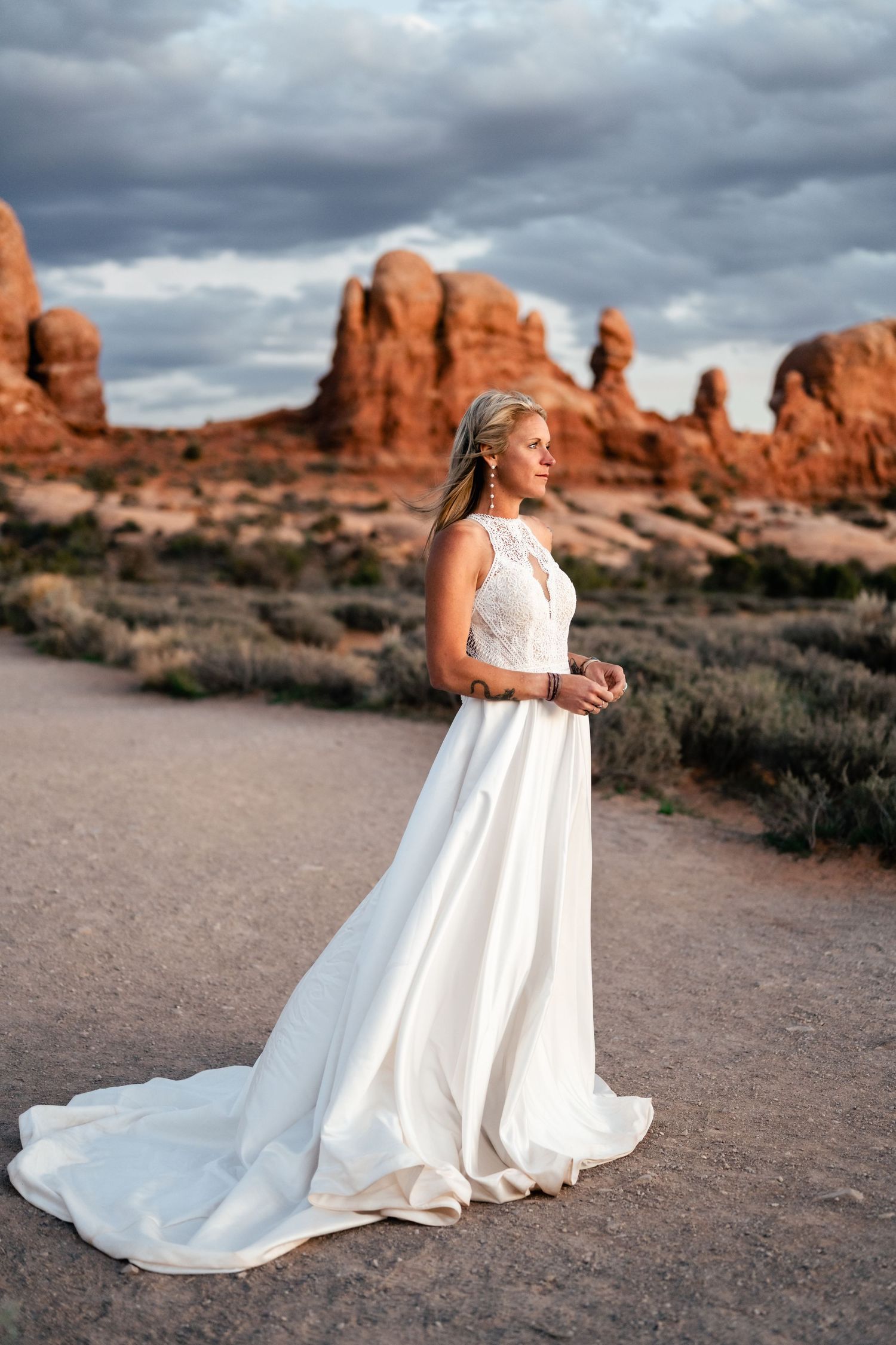 A bride in a flowing white dress poses against dramatic red rock formations in a desert landscape.