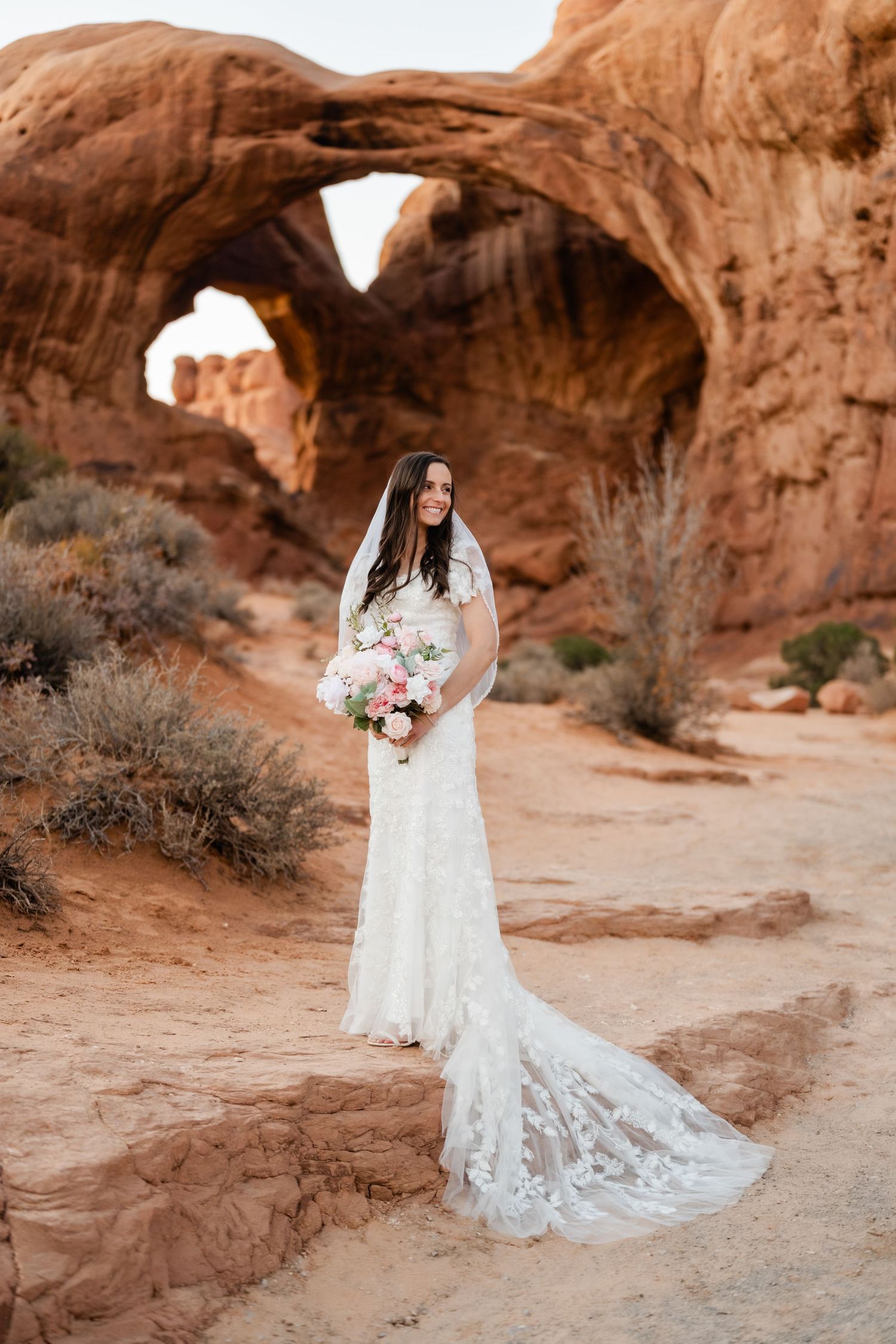 A bride in a white lace wedding dress with long train poses in front of majestic red rock arches in the desert.