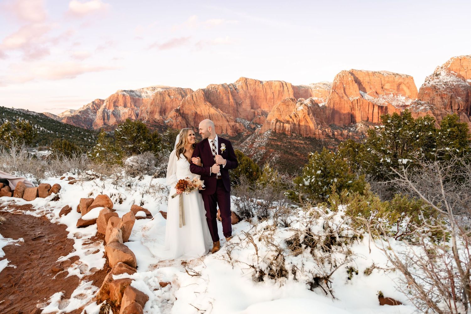 A couple in wedding attire stands on snowy terrain with majestic red rock cliffs of Zion National Park at sunset.