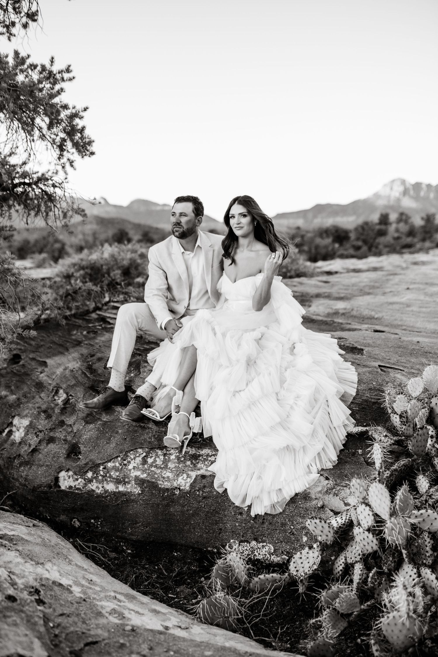 Black and white photo of a couple in wedding attire sitting on desert rocks surrounded by natural vegetation.