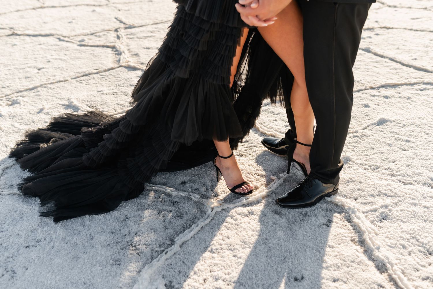 A close-up shot of elegant black and gold high heels worn with a flowing black dress on a light concrete surface. Salt Flats Photographer.