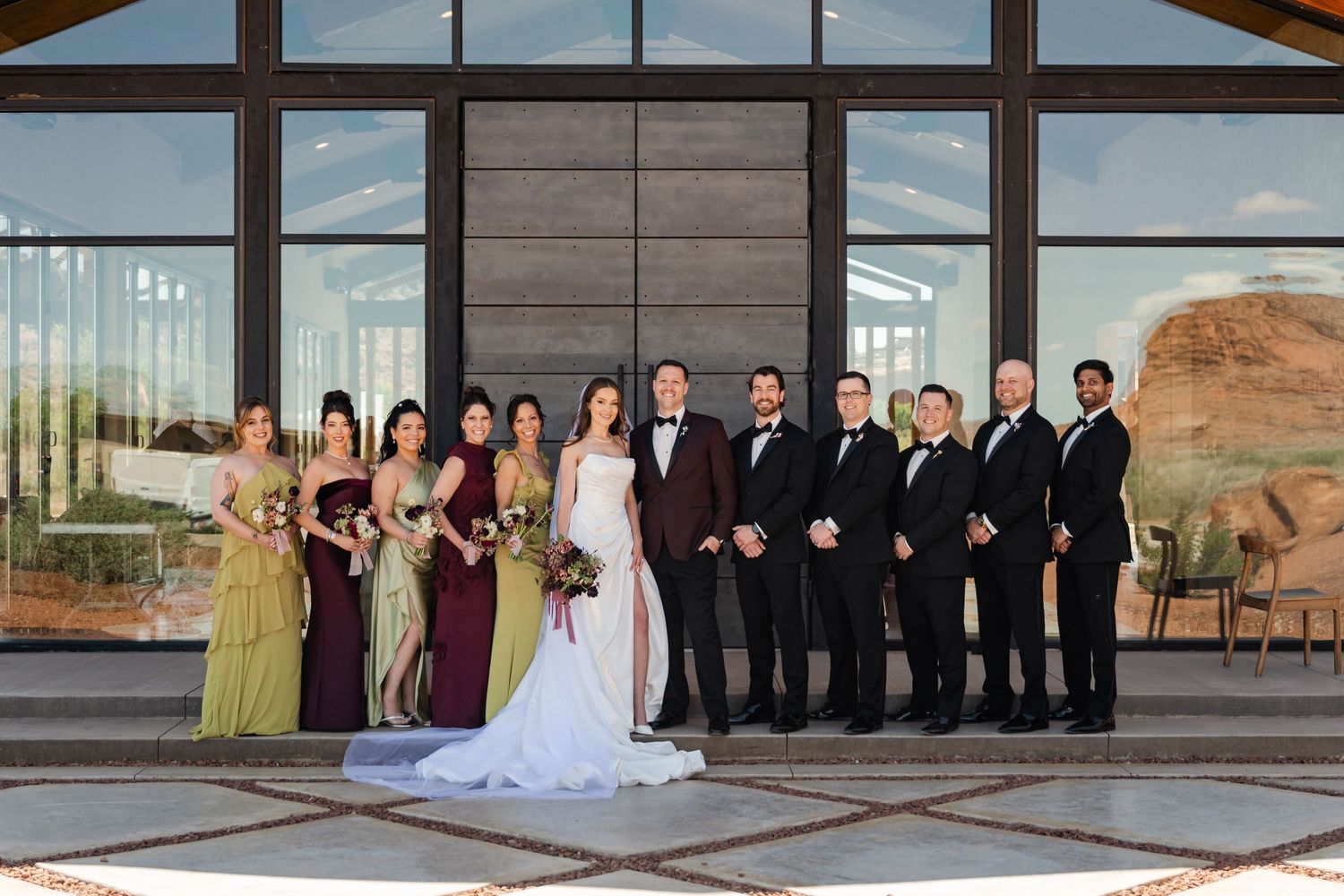 Wedding party in formal attire stands before modern venue with desert canyon views through large windows in Moab, Utah