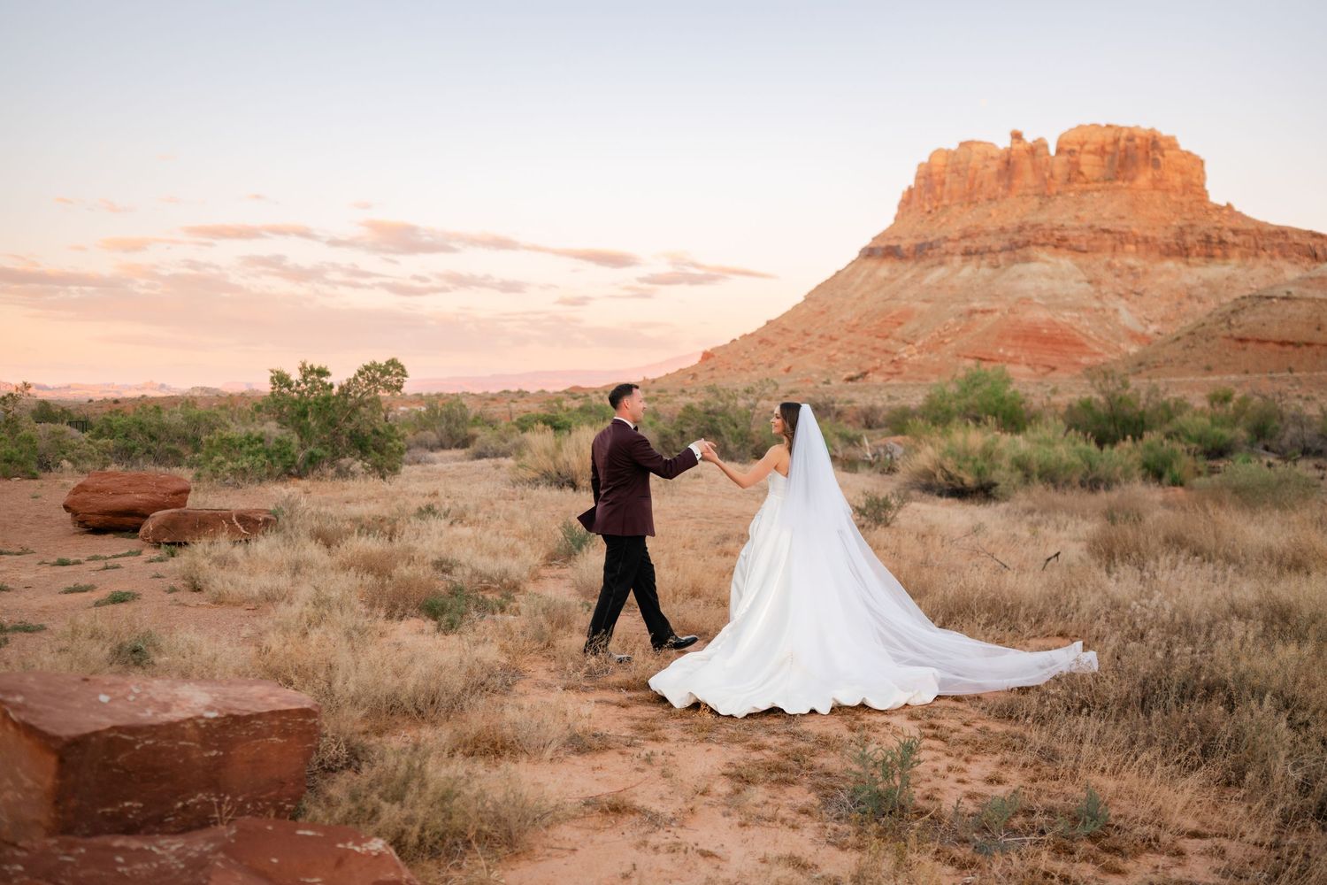 Bride and groom hold hands walking through red rock desert landscape at sunset in Utah.