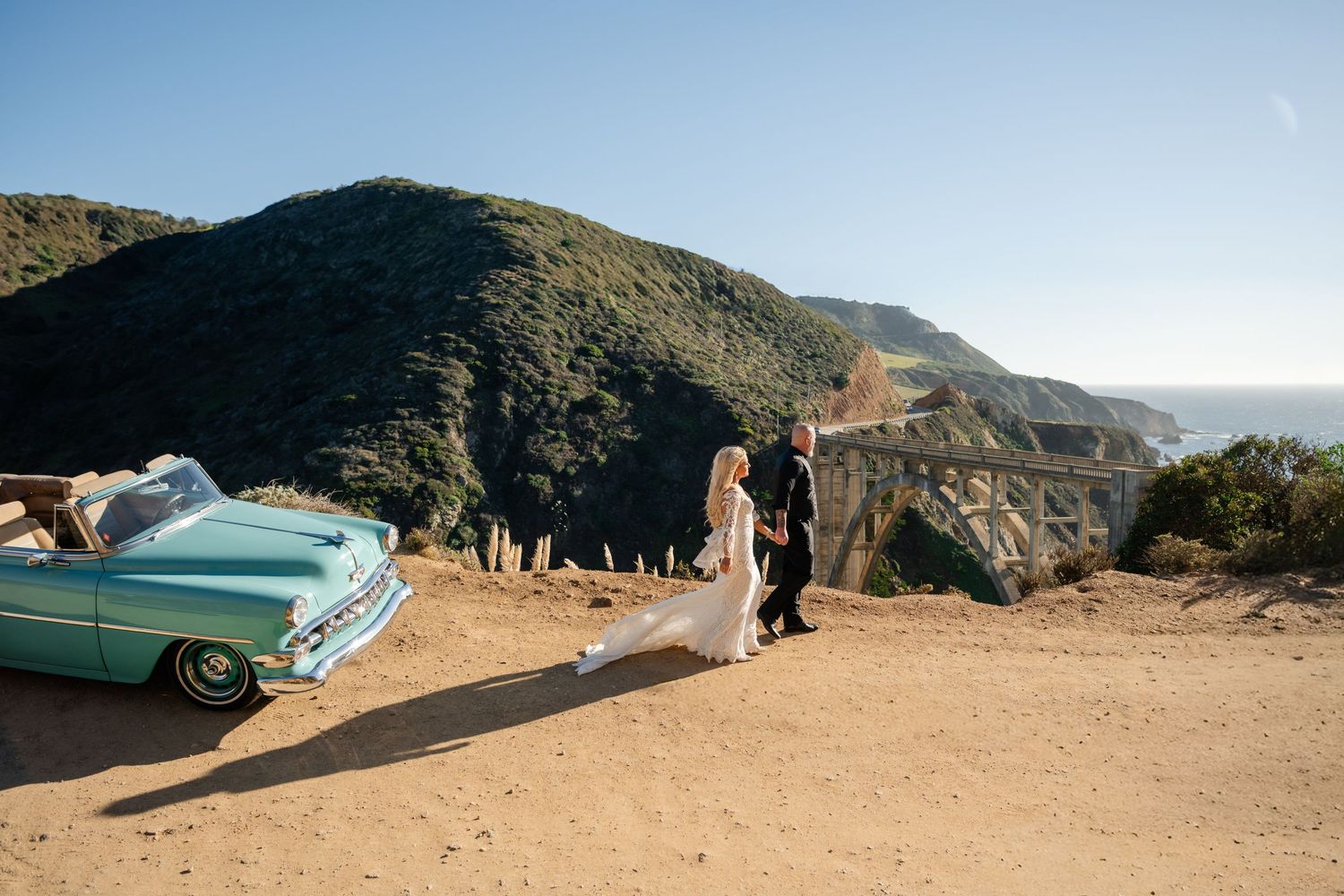 Panoramic coastal mountain views with vintage car silhouetted against blue sky.