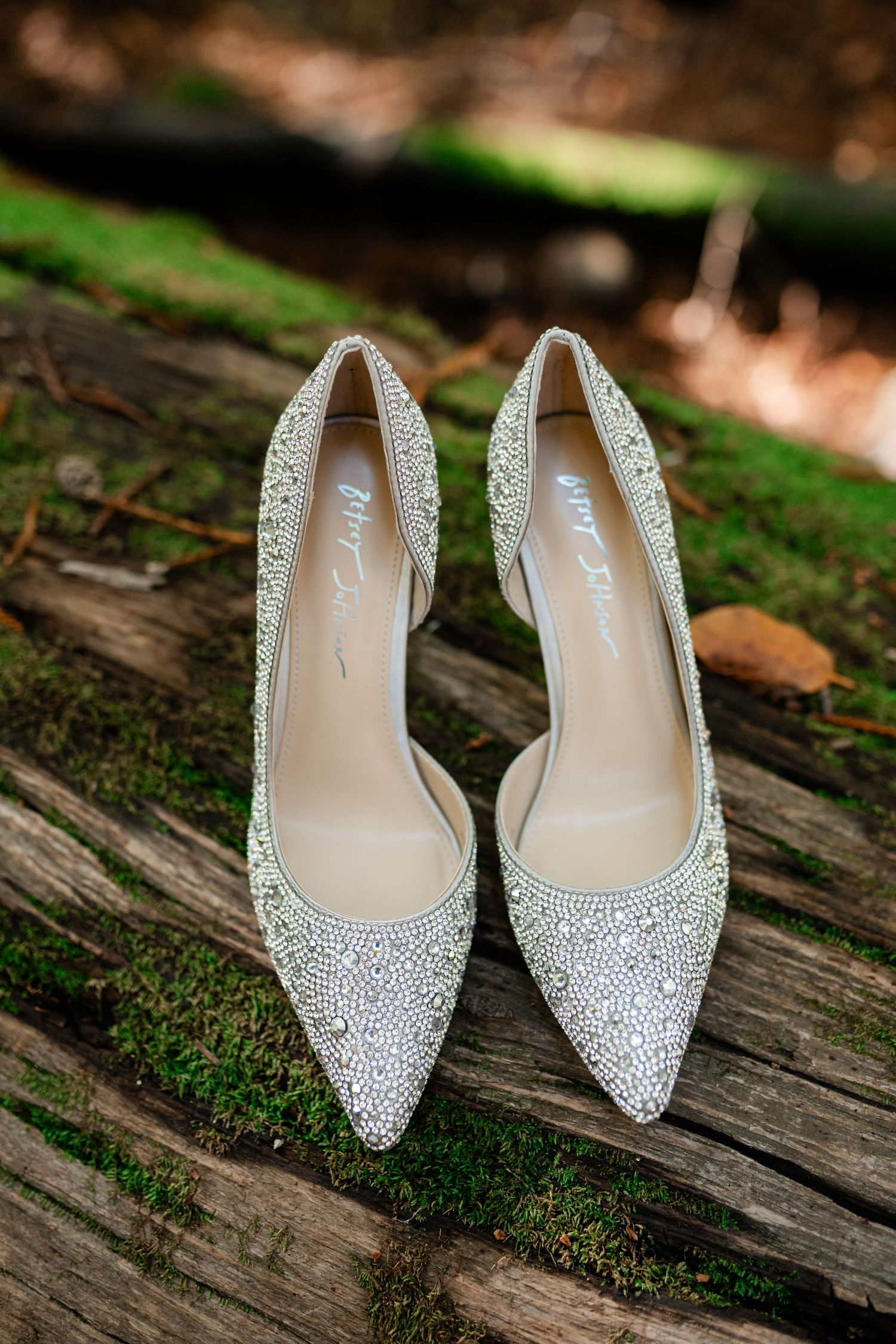 Sparkling rhinestone-encrusted wedding heels displayed on rustic wooden surface with moss details.