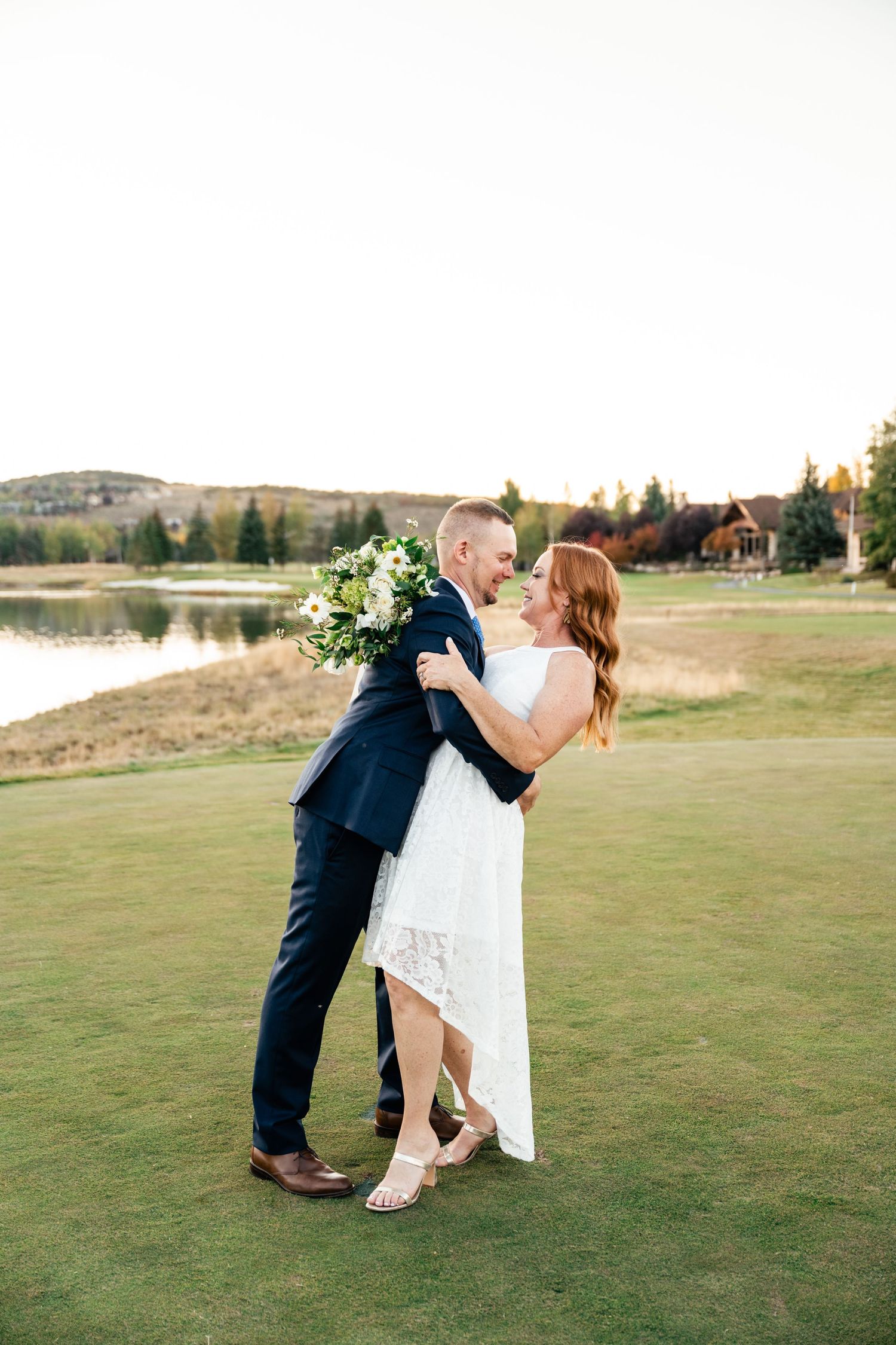A romantic wedding couple embraces on a golf course near a lake during golden hour.
