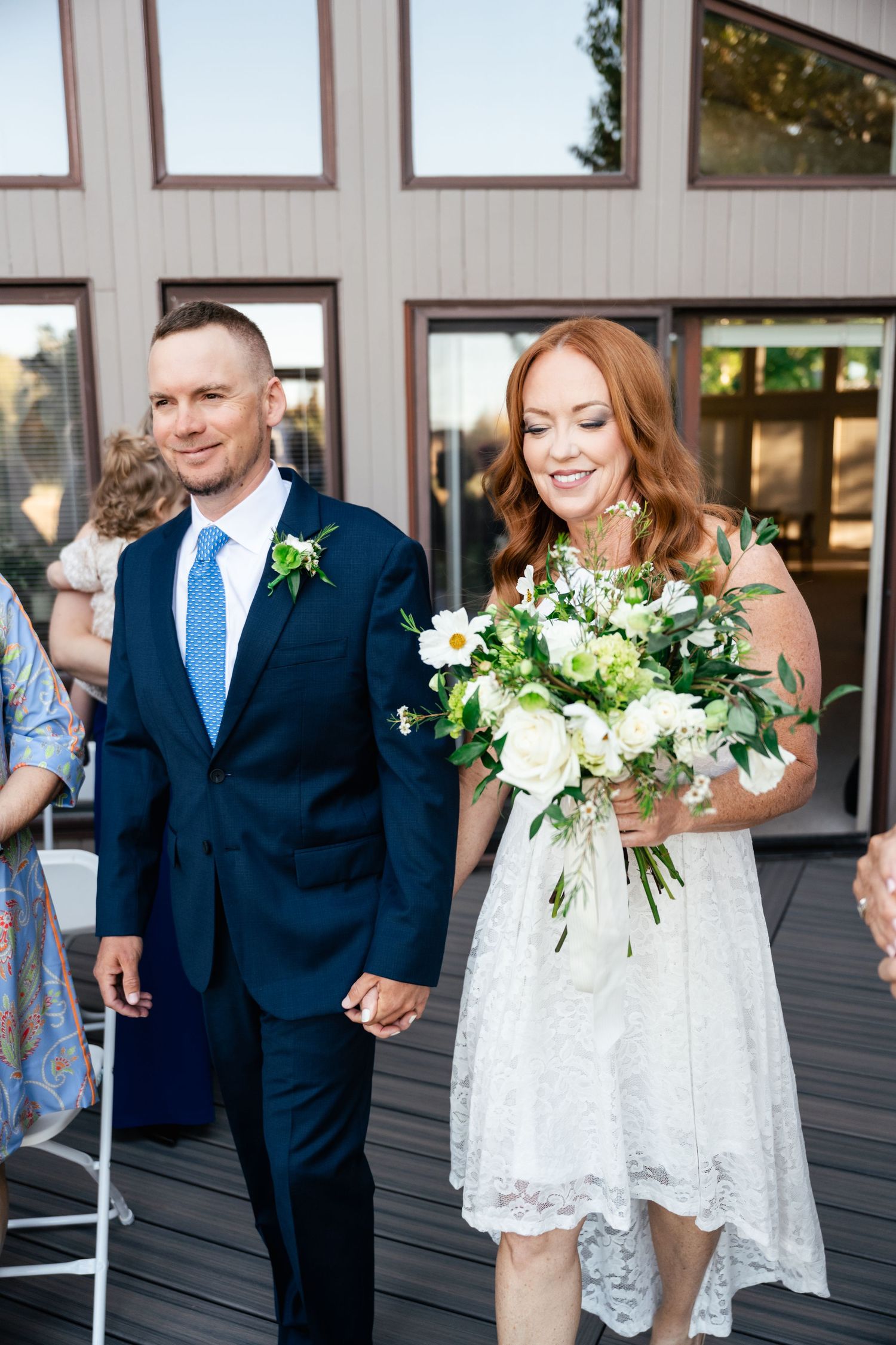 A couple holds hands while standing on an outdoor deck in formal wedding attire.
