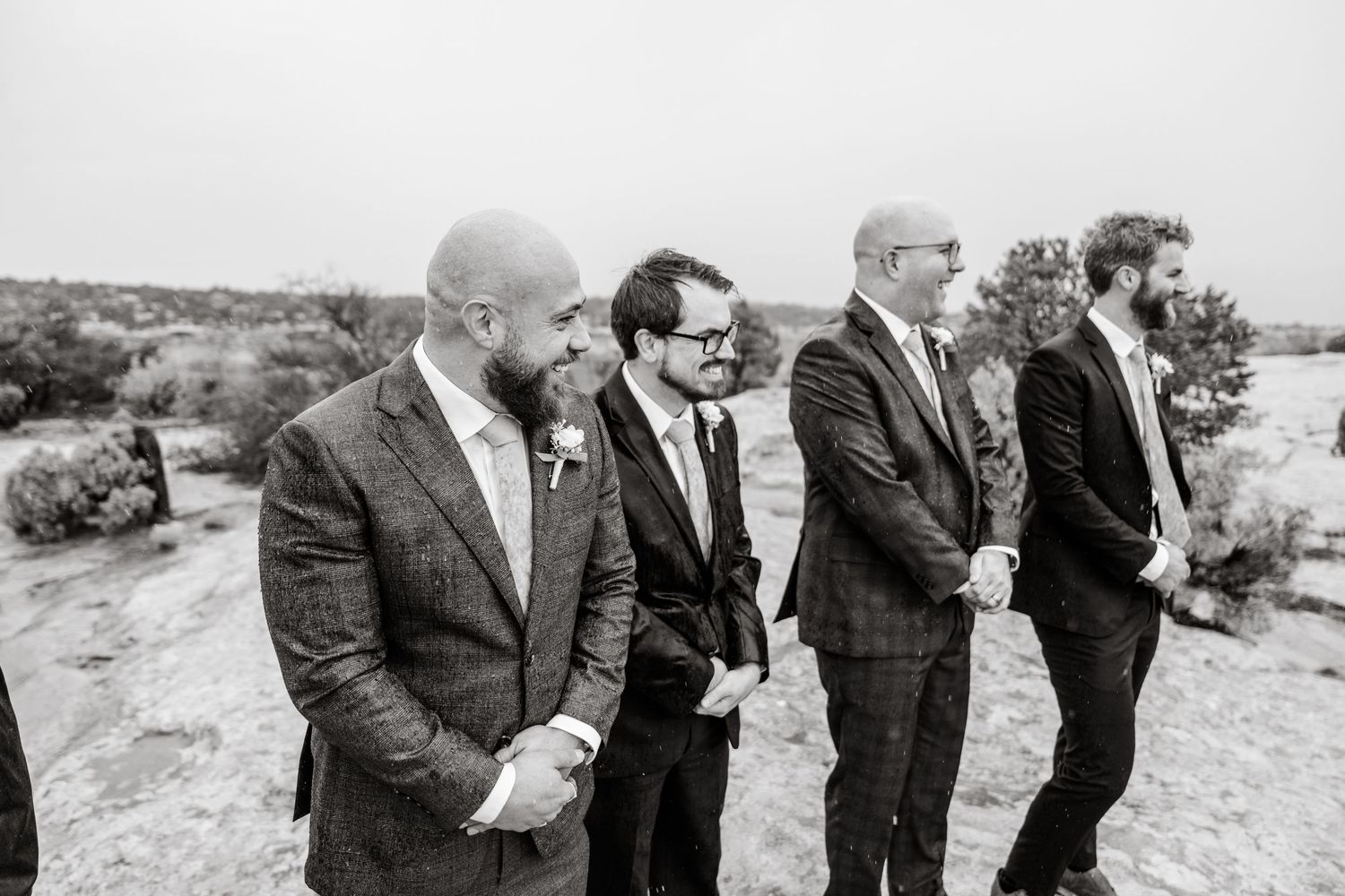 Wedding party members in formal attire socialize on desert terrain.