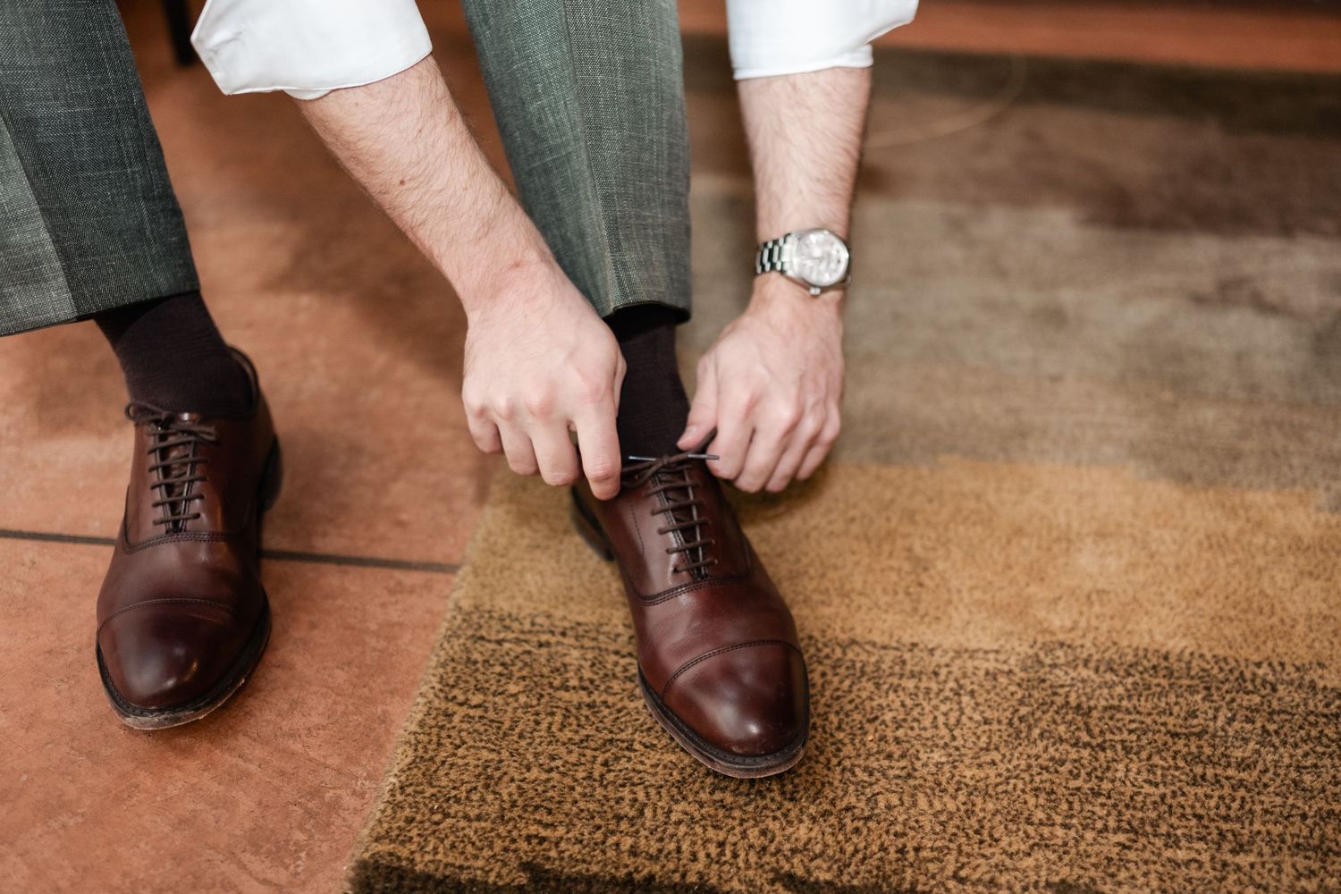 Person wearing brown leather dress shoes stands on a textured tan doormat while adjusting their footwear.