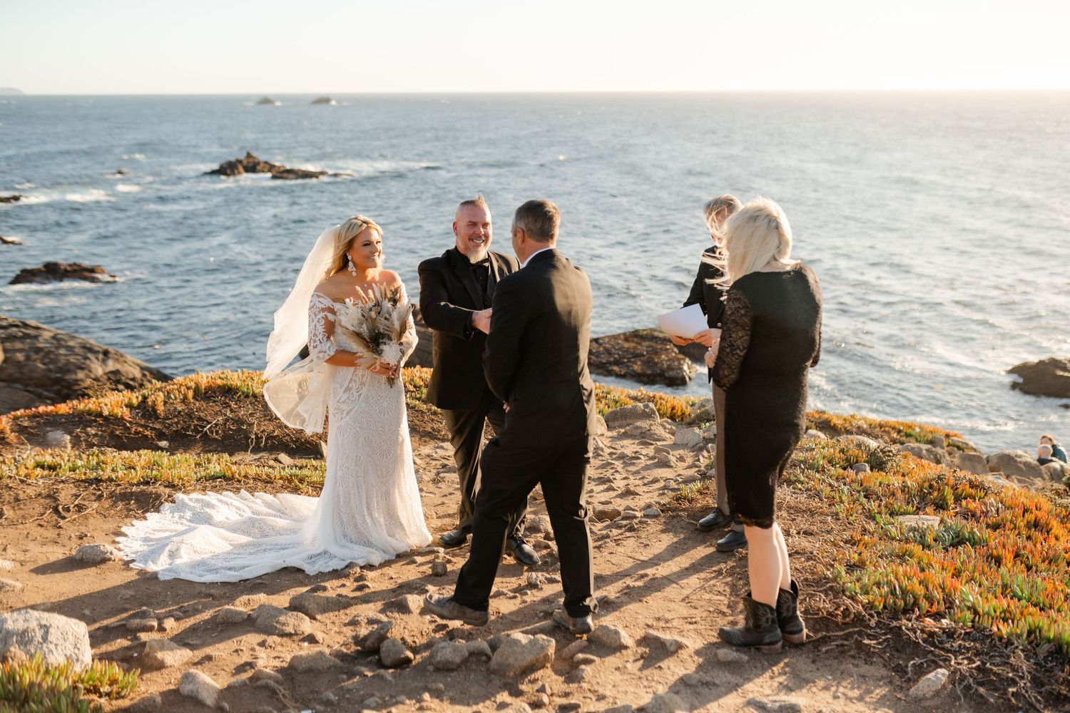 A wedding party stands together on a rocky oceanside cliff during a sunset ceremony.