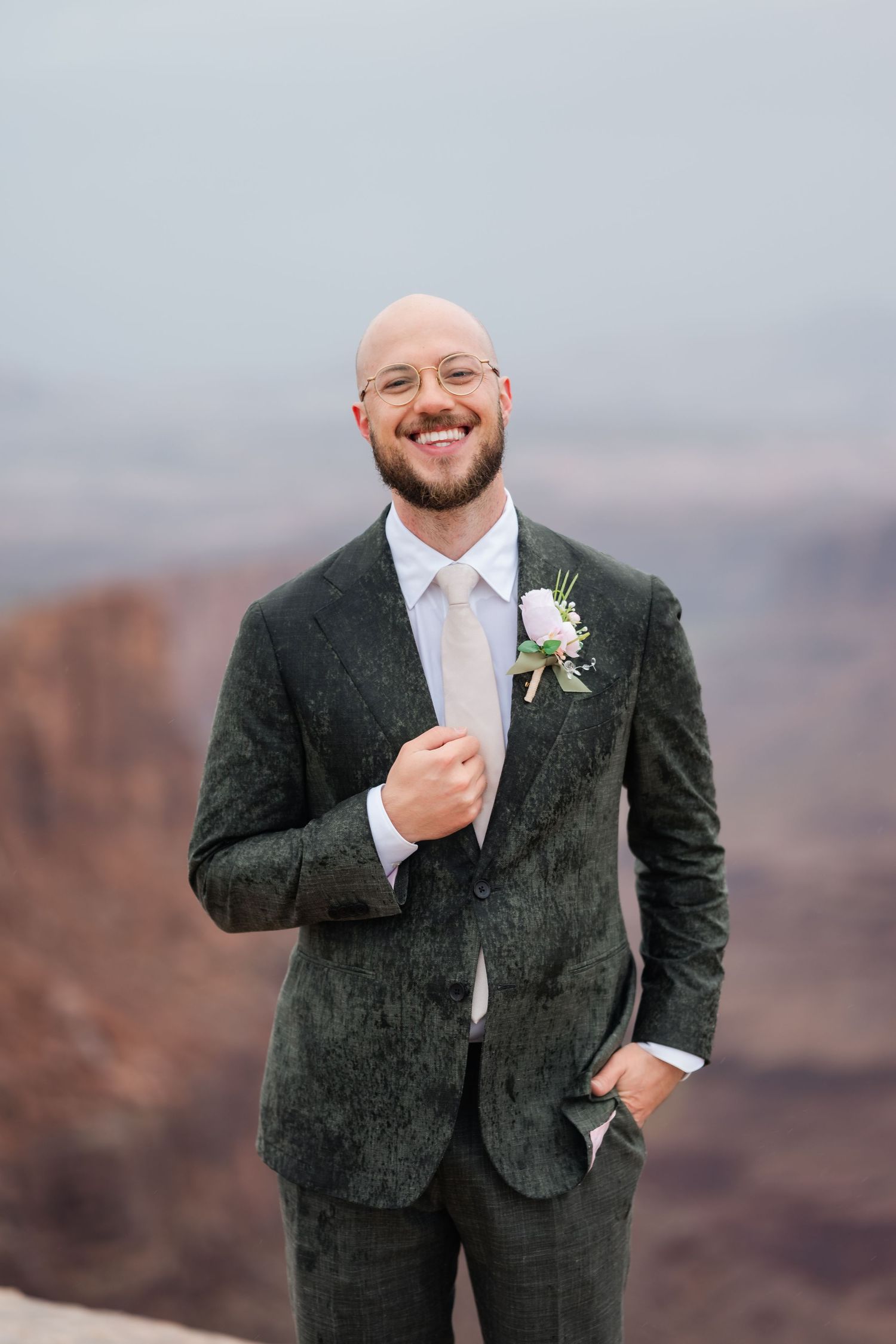 A groom in a dark suit and white boutonniere poses joyfully against a desert canyon backdrop.