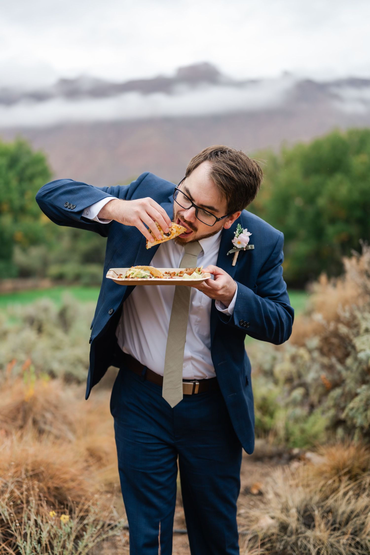 Groom in navy suit bending down dramatically in outdoor desert setting.