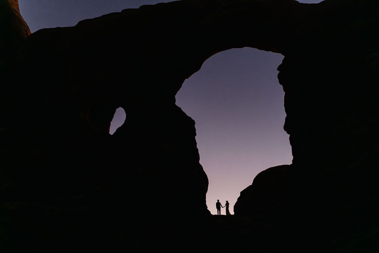 Silhouetted rock formations at dusk create a dramatic natural arch against purple sky.