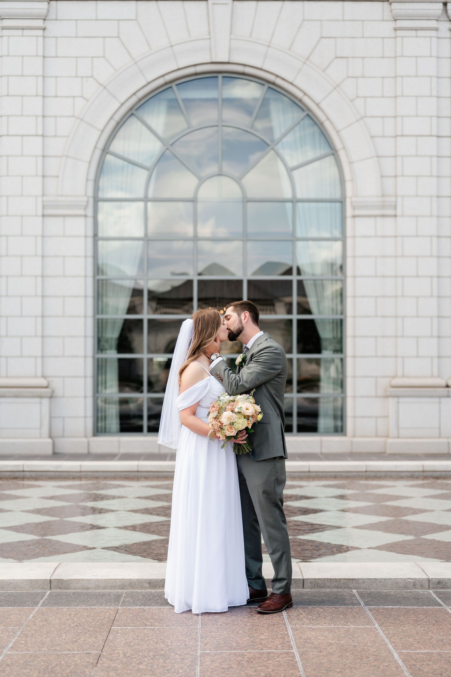 A tender moment between newlyweds against elegant white stone architecture with arched windows.