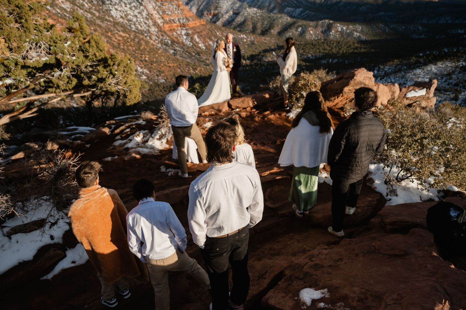 Group gathering at scenic overlook with dramatic red rock cliffs and winter snow in background.