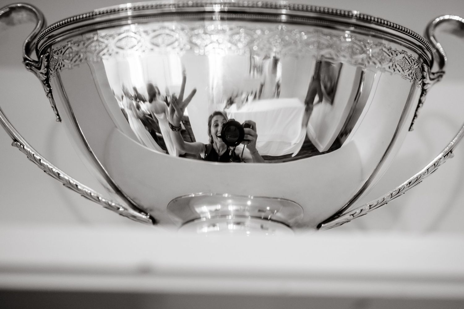 Reflective silver punch bowl with ornate handles showing distorted reflection of wedding celebration.