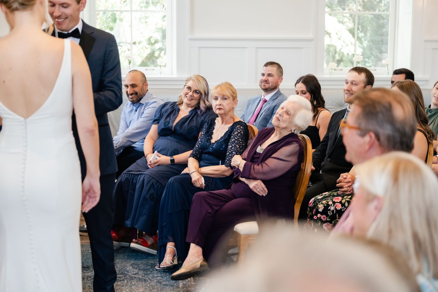 Wedding guests seated in rows laugh and react emotionally during an indoor ceremony.