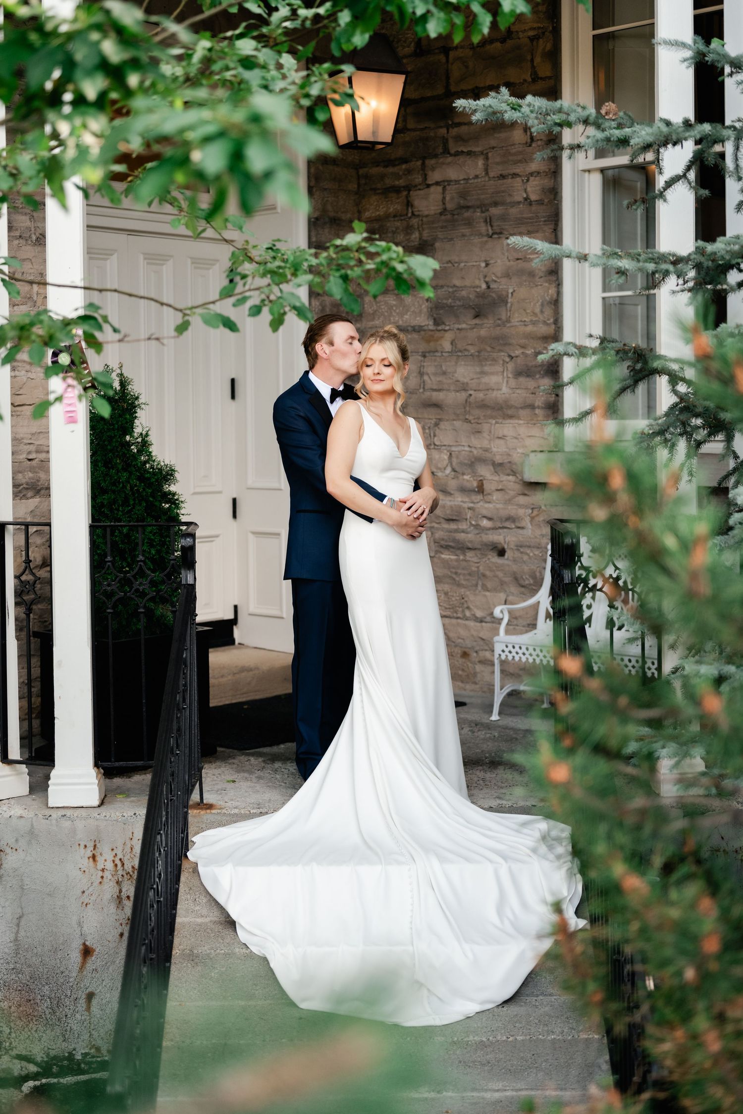 Wedding couple dances on stone porch with white columns and green foliage framing the scene.