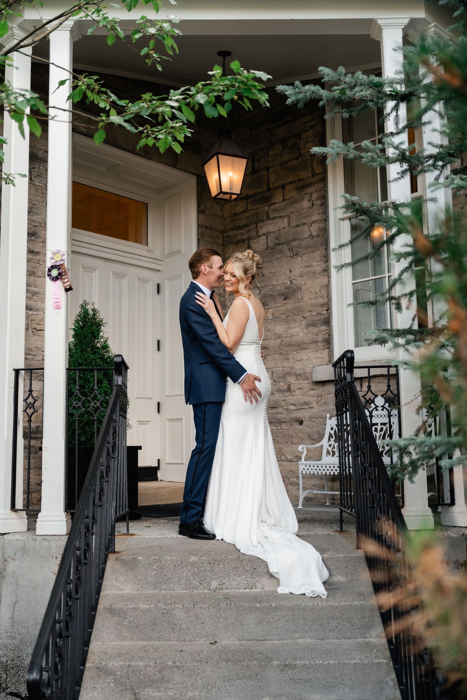 A romantic wedding couple shares an intimate moment on a stone porch with white columns and greenery.