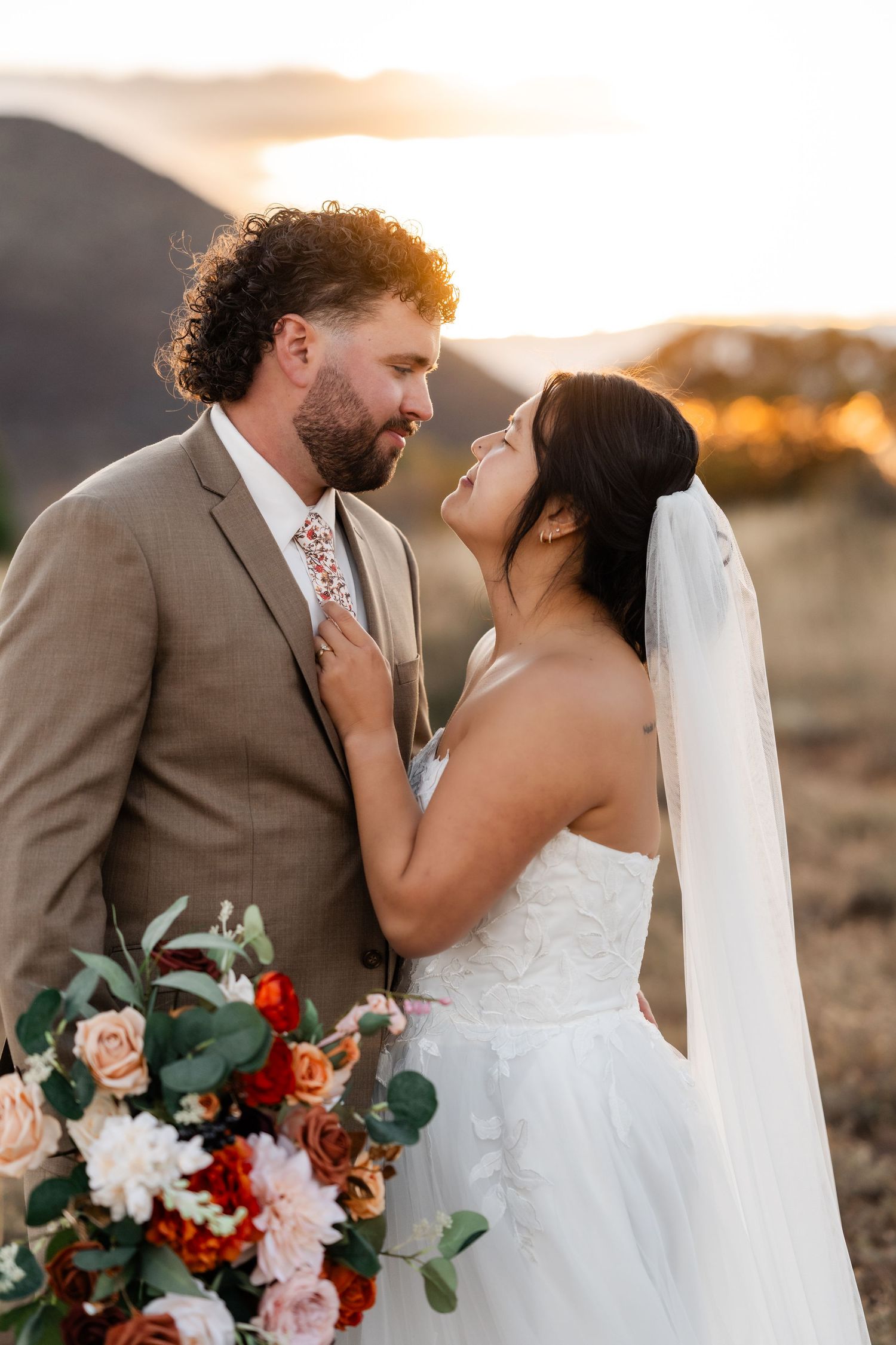 A wedding couple embraces while holding a bouquet of red and white flowers at sunset.