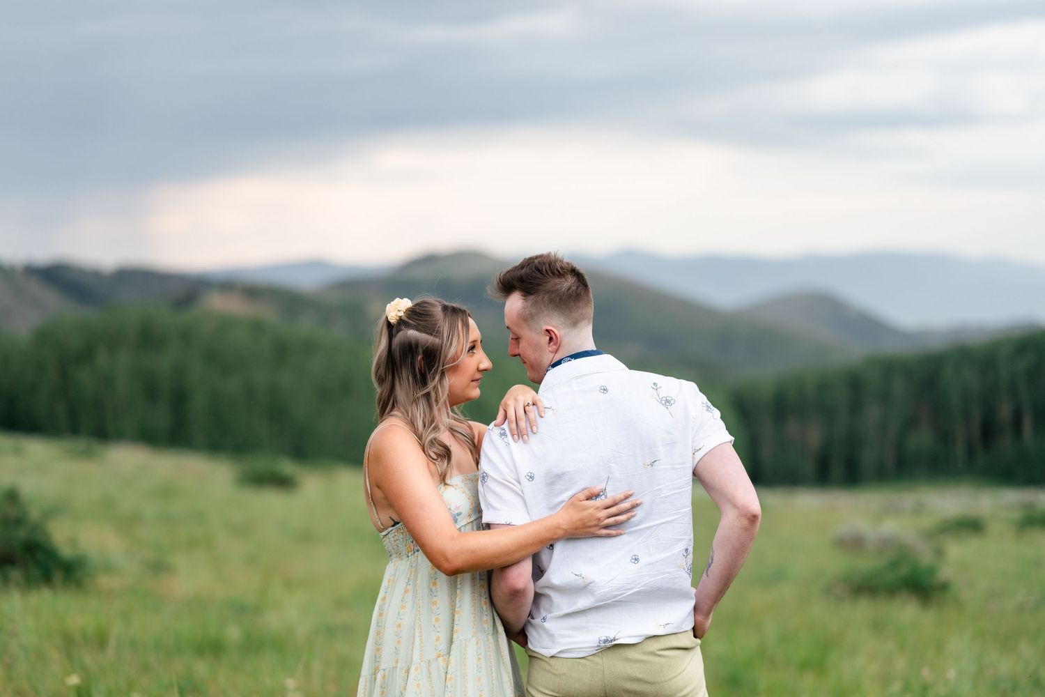 A couple embraces in a mountain meadow with dramatic peaks and forests in the distance.