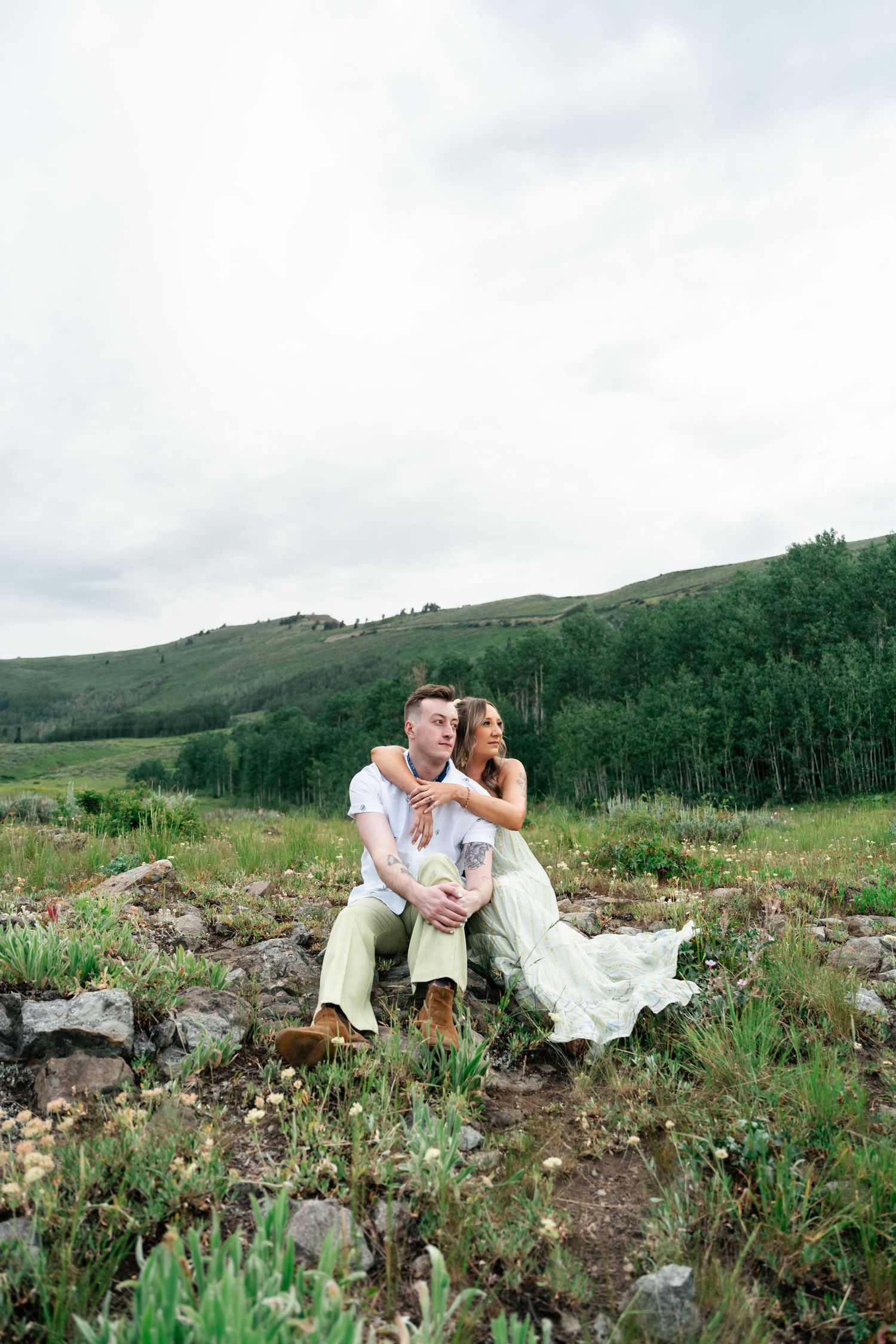 A couple sits together on rocky terrain surrounded by mountain wildflowers and aspen trees in the background.