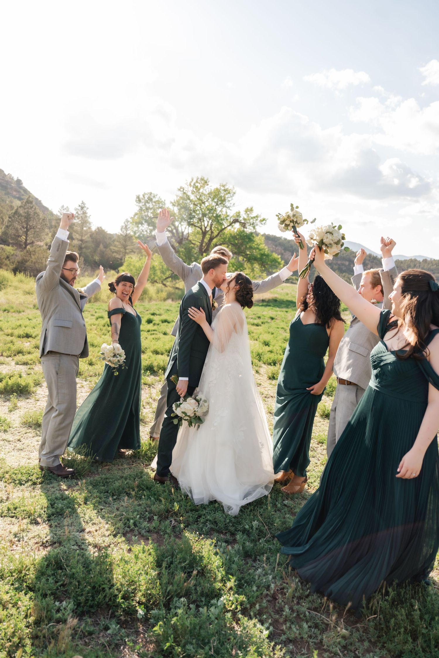 Wedding party celebrates joyfully outdoors with raised arms against mountain backdrop.