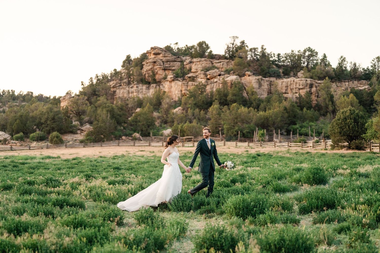 Newlyweds walk hand in hand through a green field with rocky cliffs in the background.