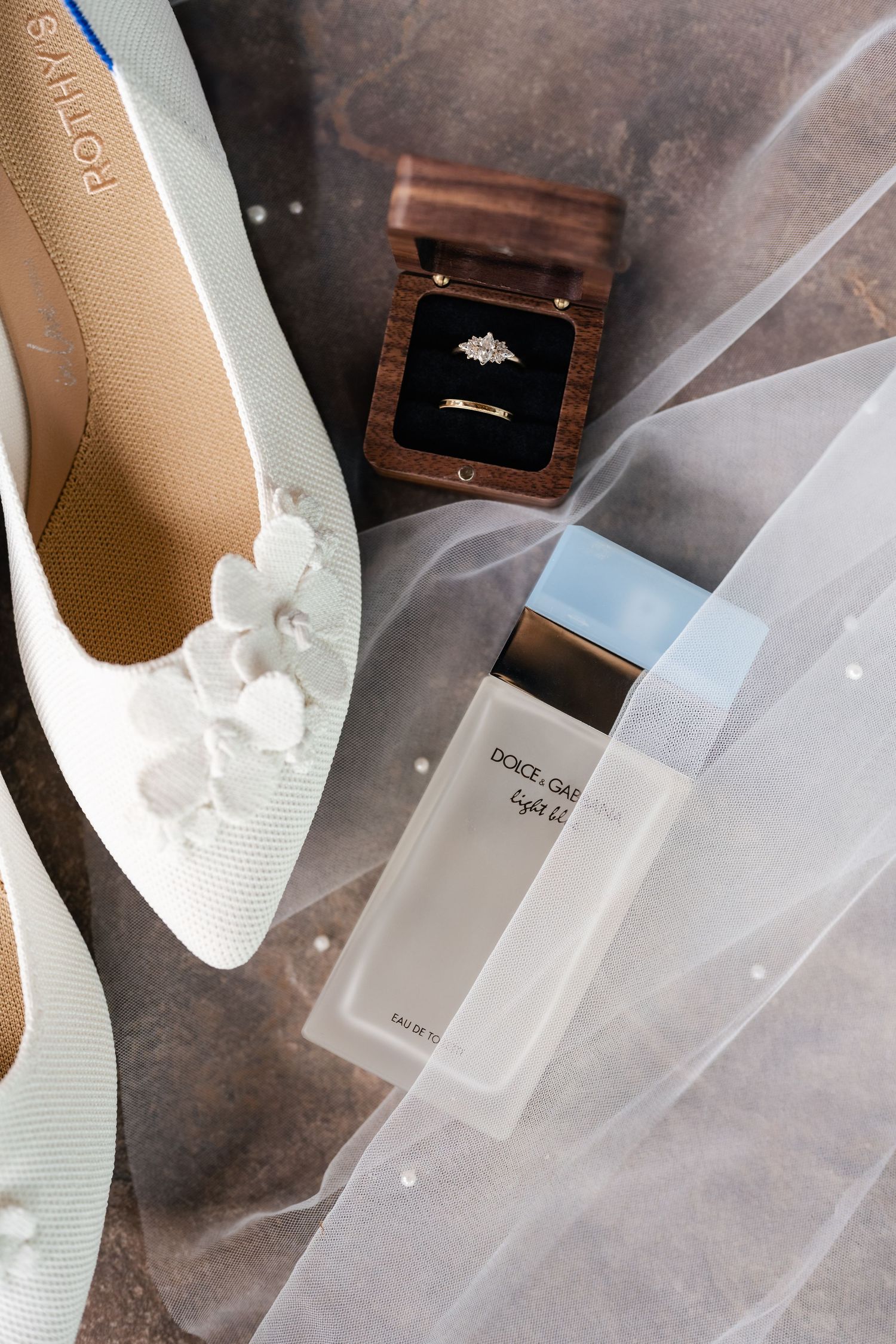 Wedding accessories including white shoes, ring box and veil arranged on textured surface.
