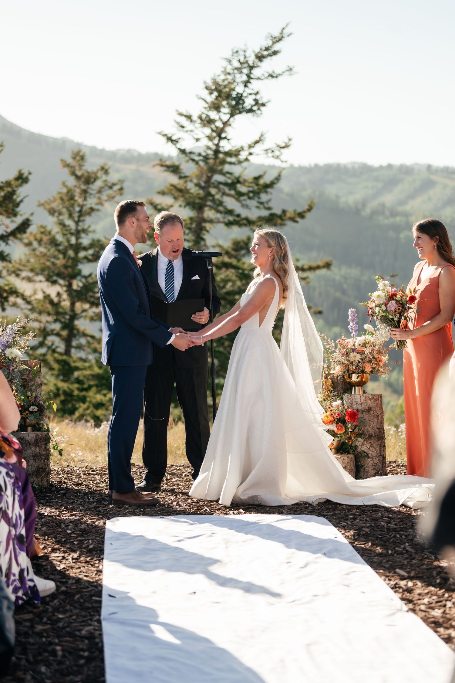 An outdoor mountain wedding ceremony with white aisle runner and evergreen trees in the background.
