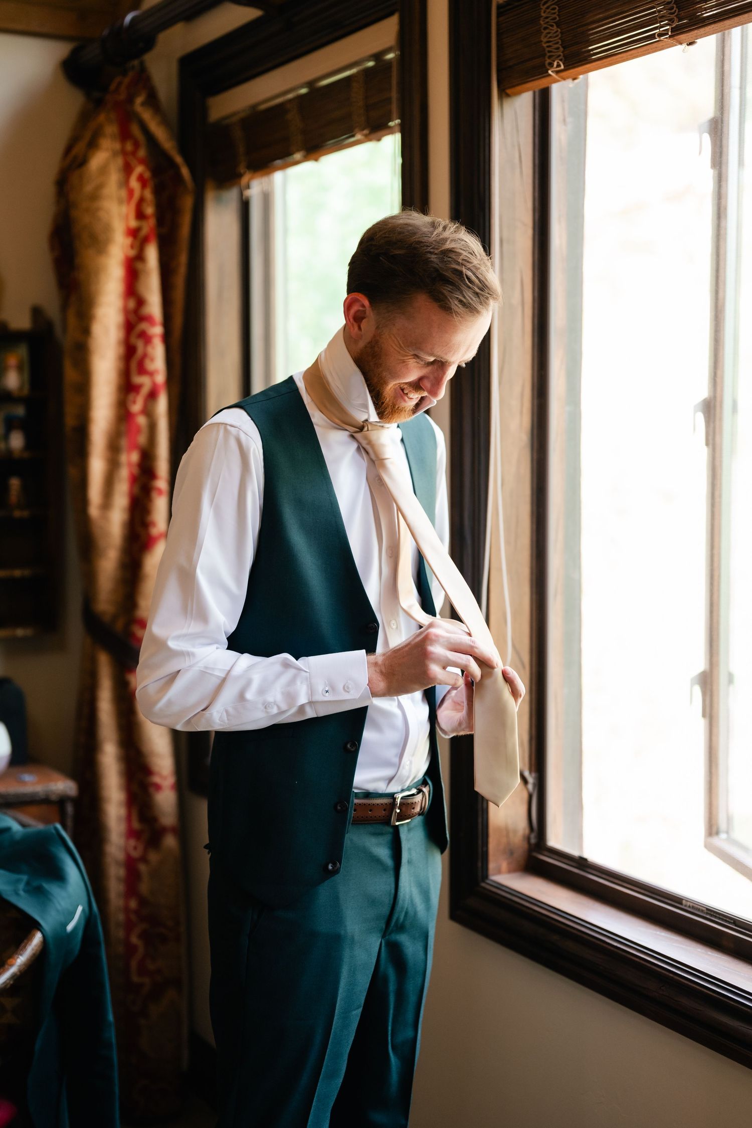 Person in formal vest and white shirt adjusting attire near window with natural lighting.