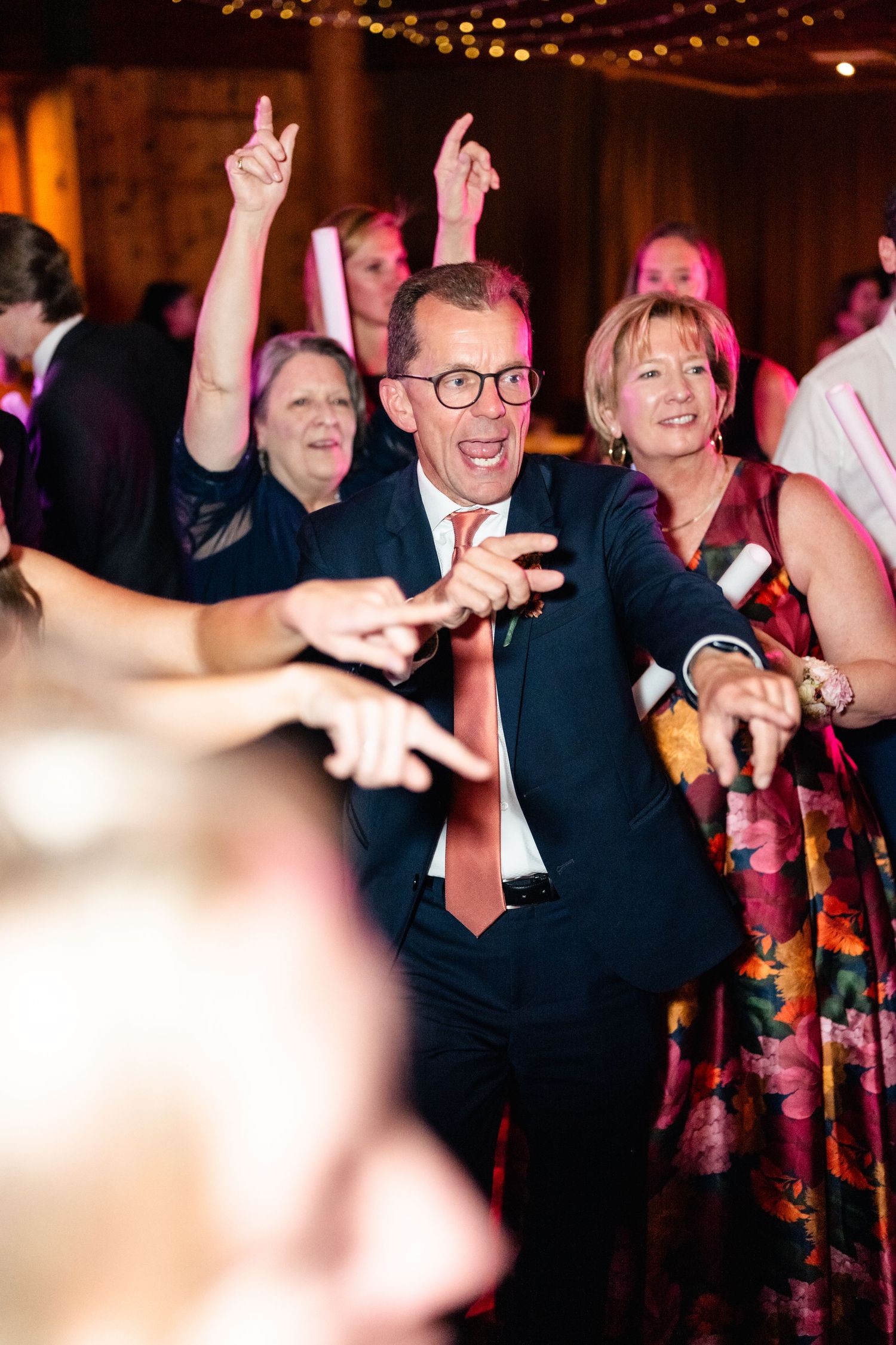 Wedding guests dancing and celebrating with raised arms under warm lighting at reception.