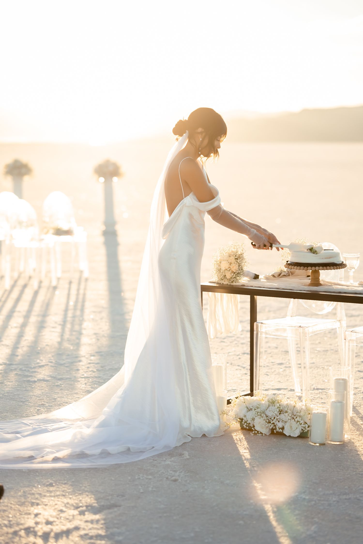 A bride in a flowing white dress cuts cake at an elegant outdoor reception table at sunset.