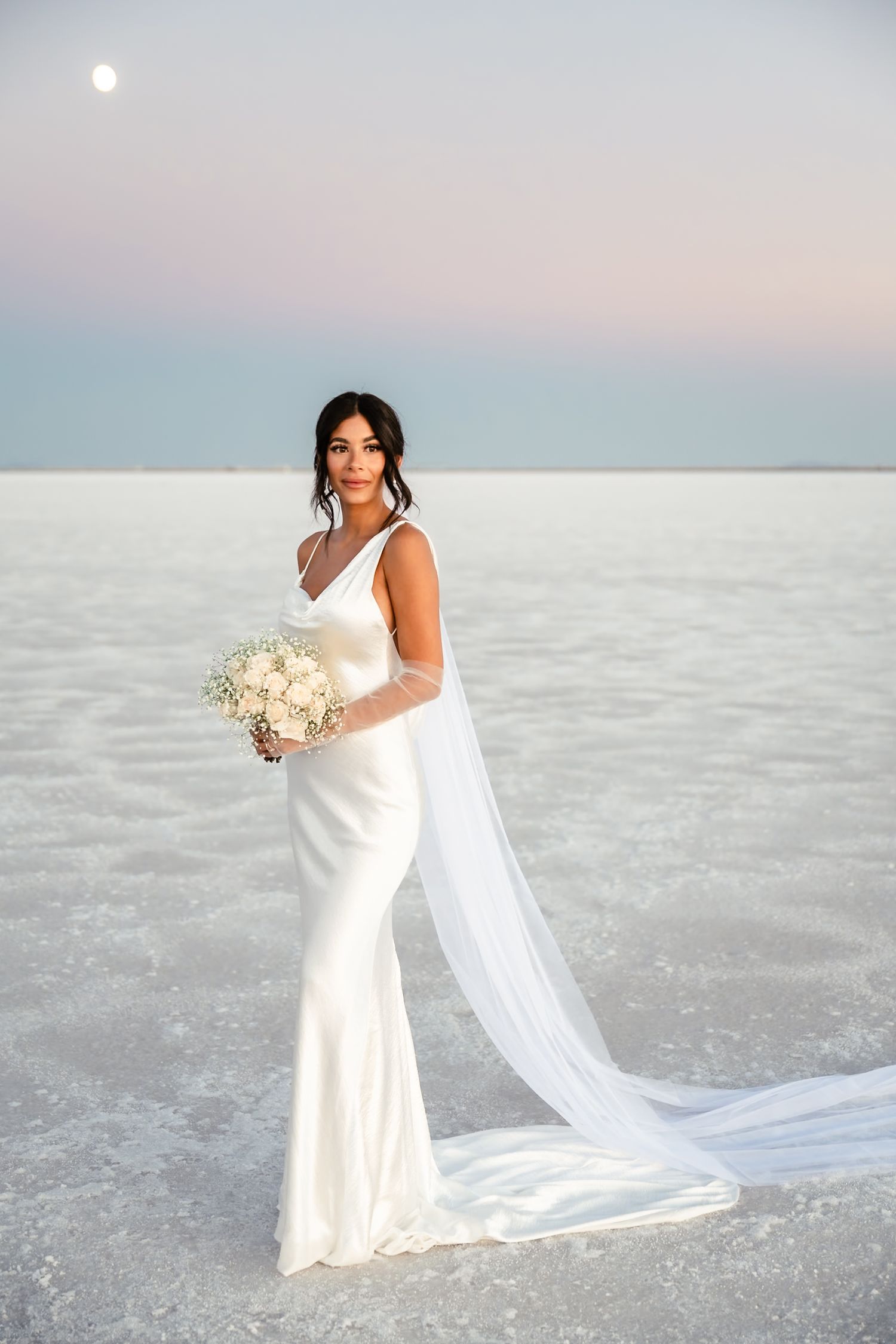 Bride in flowing white dress poses elegantly on salt flats at sunset.