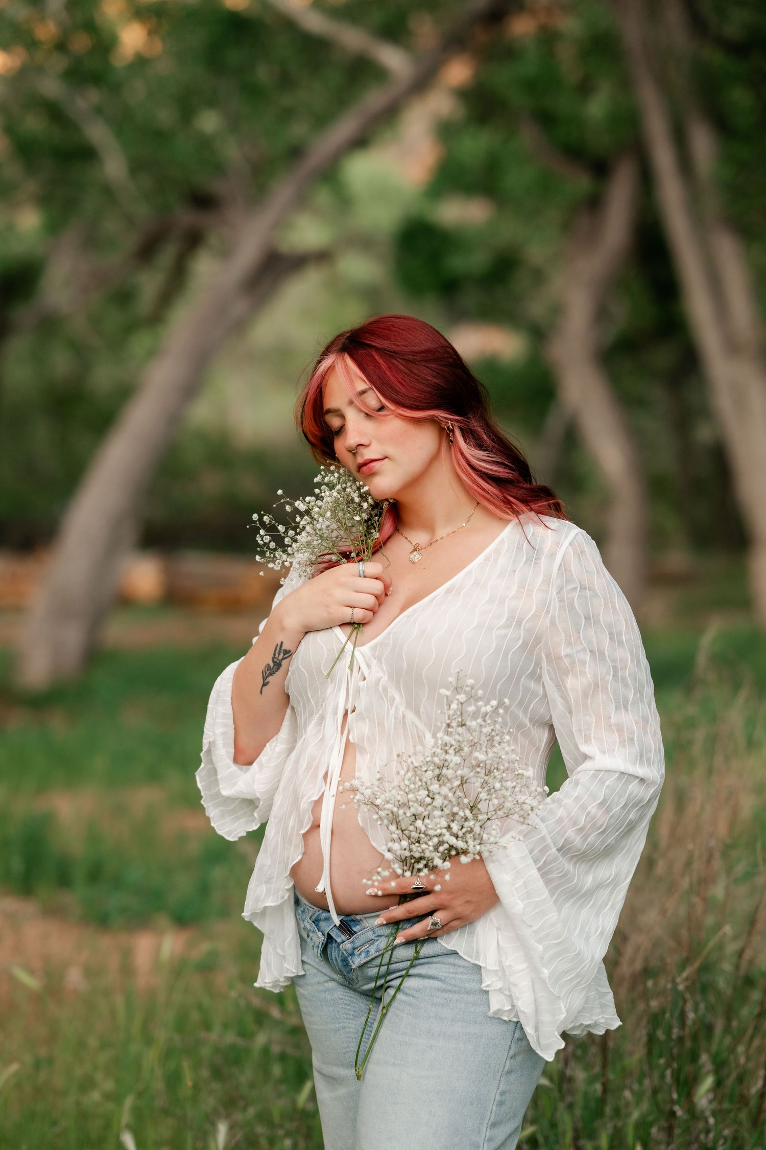 Pregnant woman with red hair in white blouse holding baby's breath flowers outdoors in a lush green park setting.