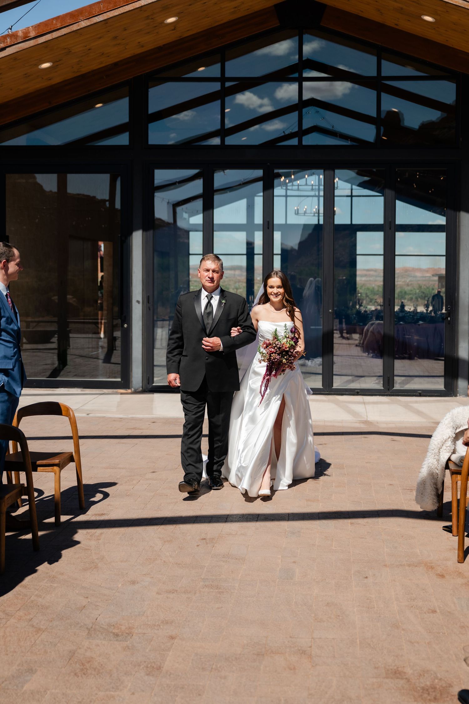 Bride walks down outdoor aisle with escort, holding bouquet, before modern glass venue on sunny wedding day.
