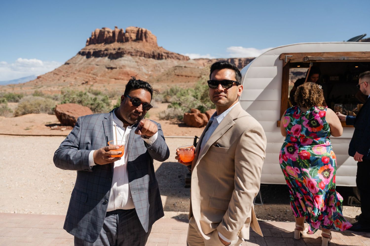 Two stylish men in suits holding drinks at an outdoor desert wedding with red rock buttes in the background.
